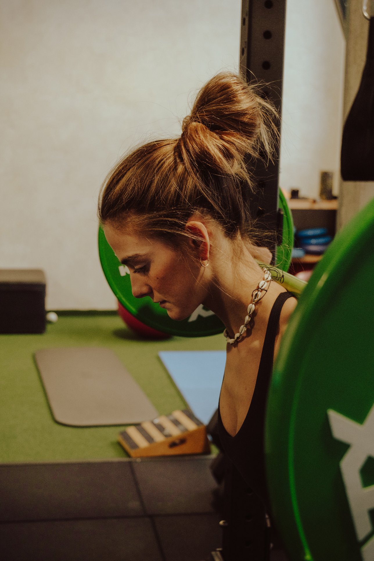 A woman with styled hair in a bun, wearing a shell necklace, sitting at a workspace with yoga mats and small percussion instruments in front of her, in a room with workout equipment.