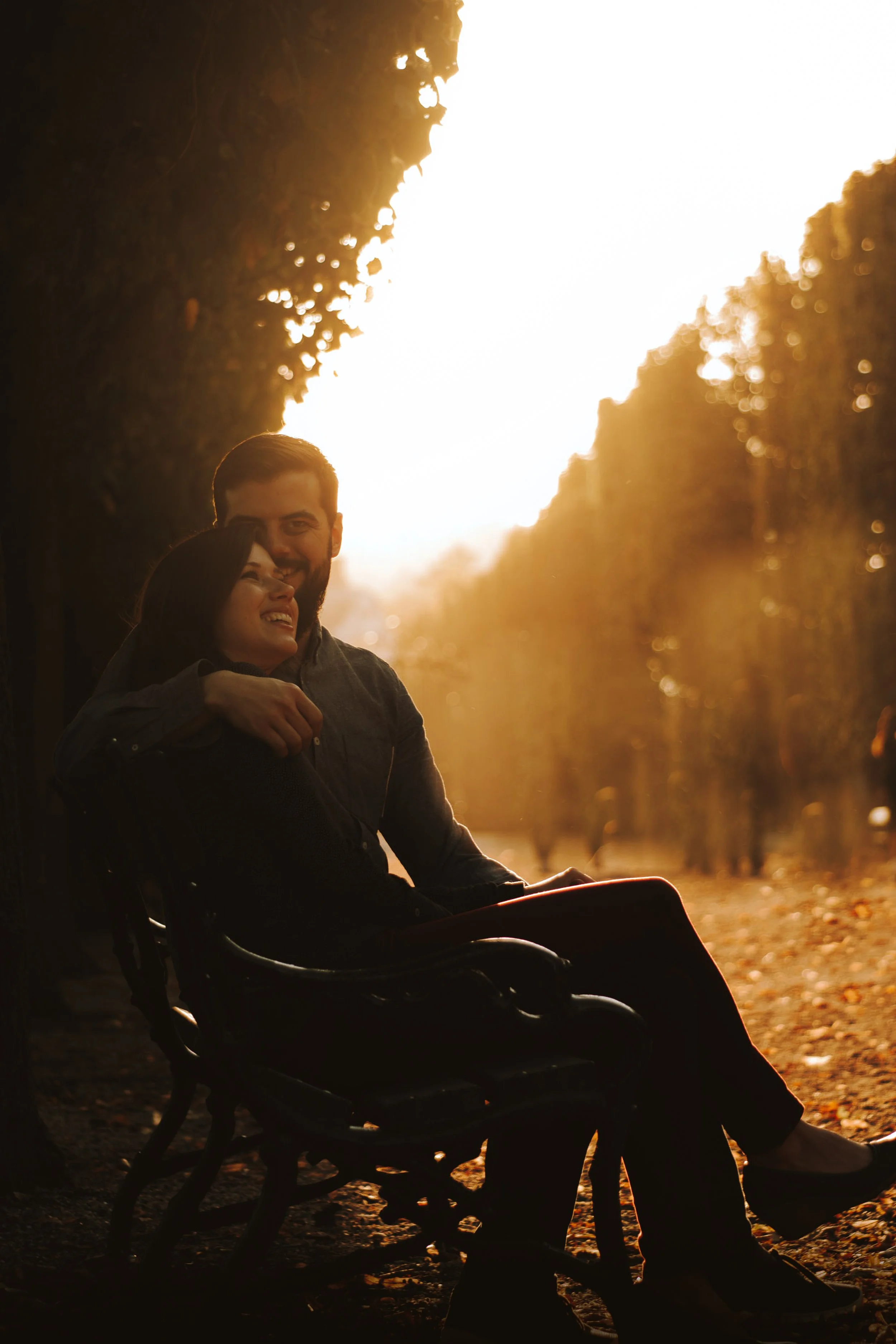 A man and a woman sitting on a bench in a park during sunset. The woman is smiling and the man has his arm around her. Trees with fall foliage line the pathway, creating a warm, golden atmosphere.
