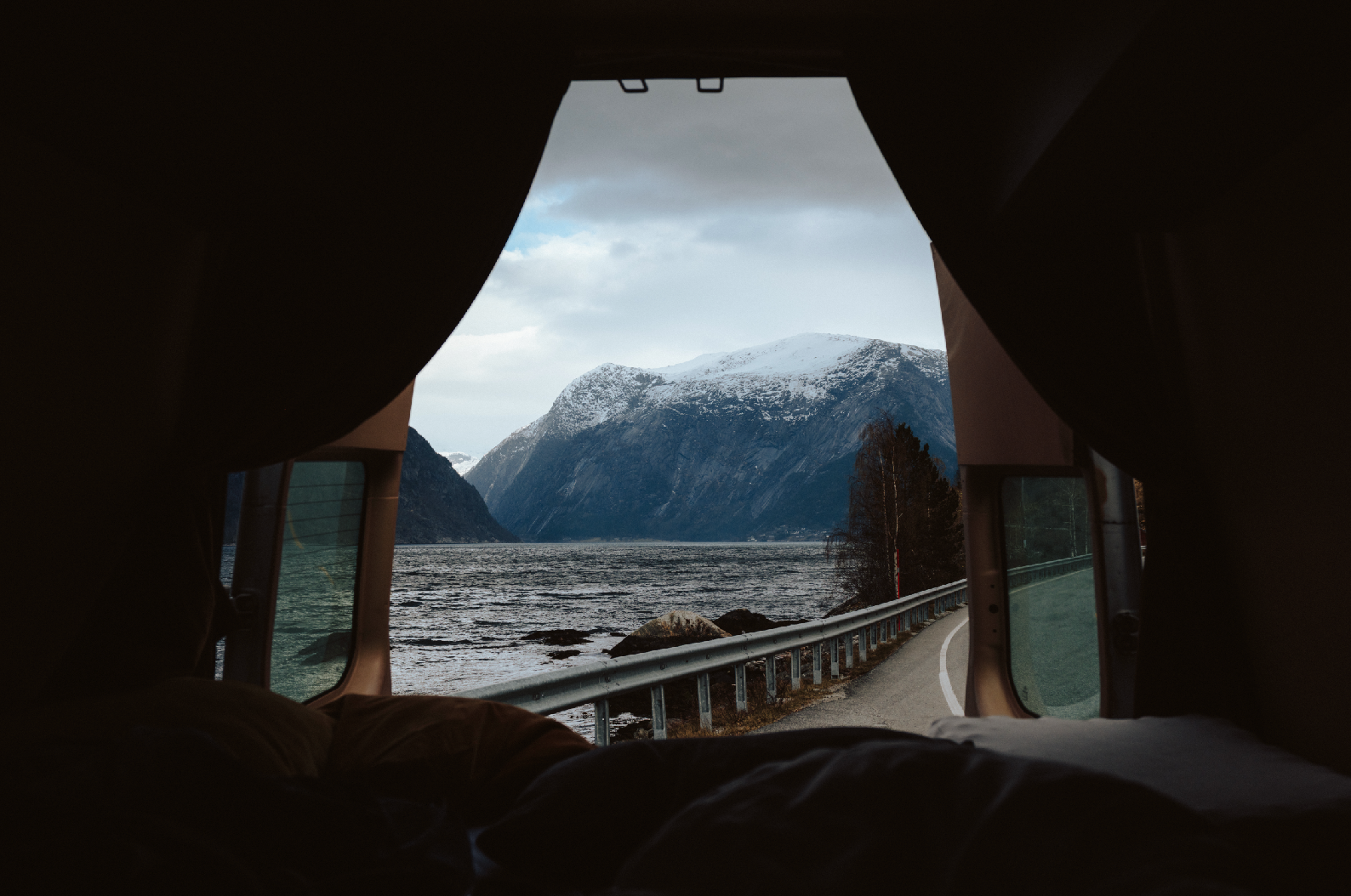 View of snow-capped mountains and a river seen through the open rear of a camper van, with a winding road along the water.
