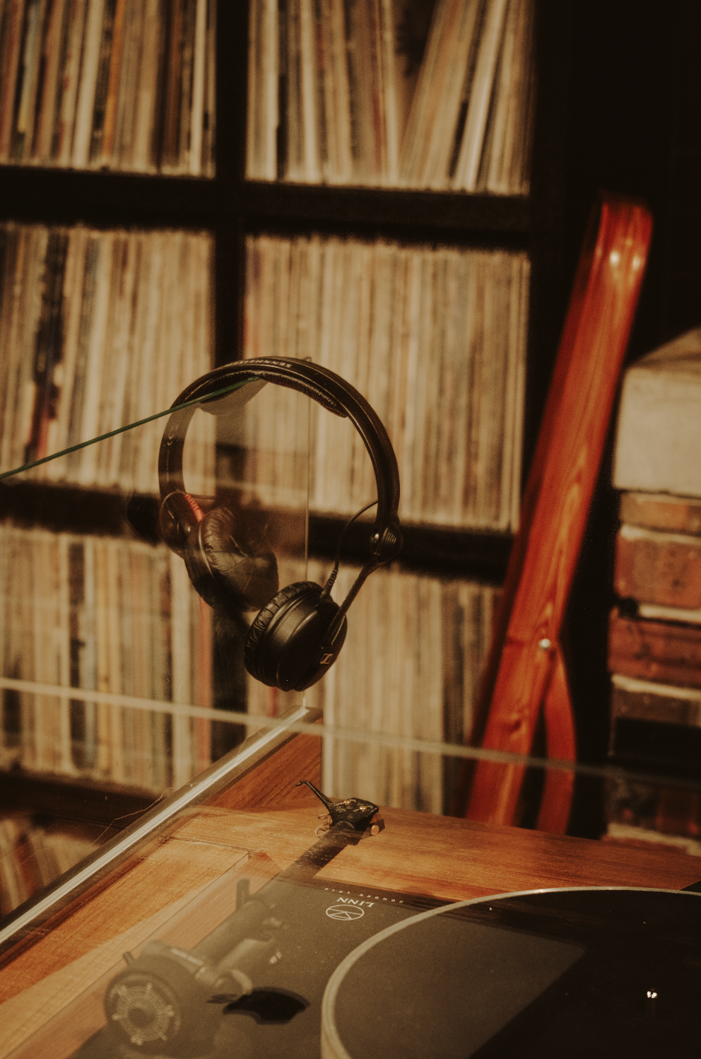 A pair of black over-ear headphones hanging on a clear glass panel, with a vinyl record collection shelf and a red wooden chair in the background.