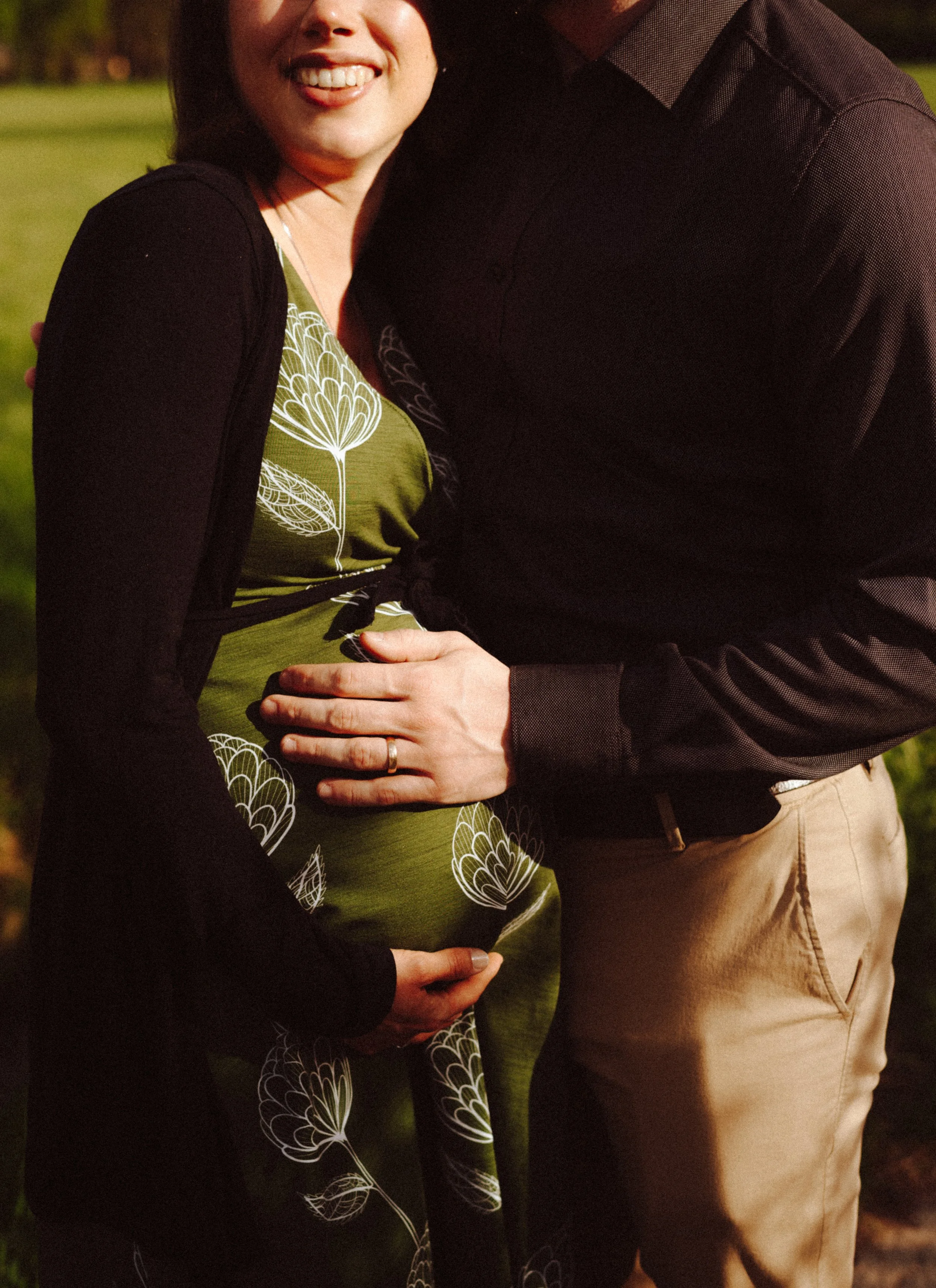 A smiling woman in a black cardigan and green dress with a white floral pattern stands close to a man wearing a dark shirt and khaki pants, both outdoors at sunset with a blurred background of greenery.