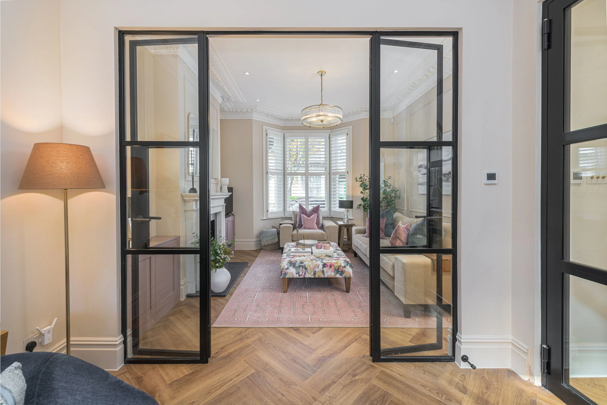 Living room viewed through black-framed glass door, with cream walls, a white fireplace, a chandelier, and large windows with white shutters, decorated with pink and gray pillows and a pink rug.