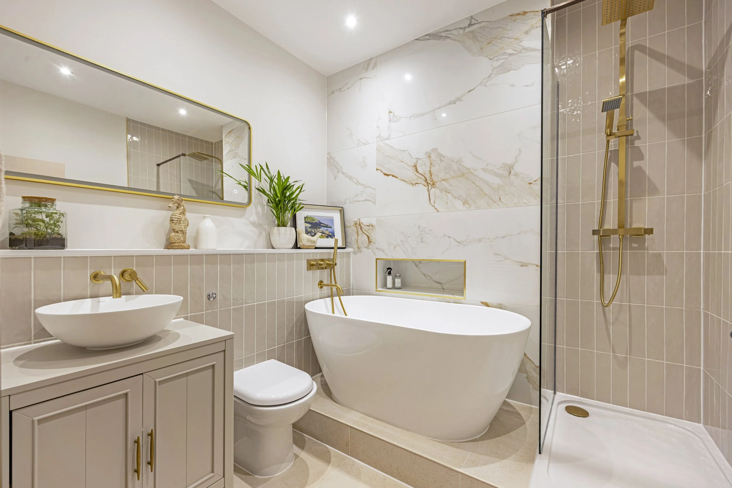 Modern bathroom featuring a freestanding bathtub, a beige vanity with a vessel sink, a large wall mirror, decorative items, a potted plant, framed artwork, beige tile walls with marble accents, a shower with a glass door, a gold shower fixture, and r