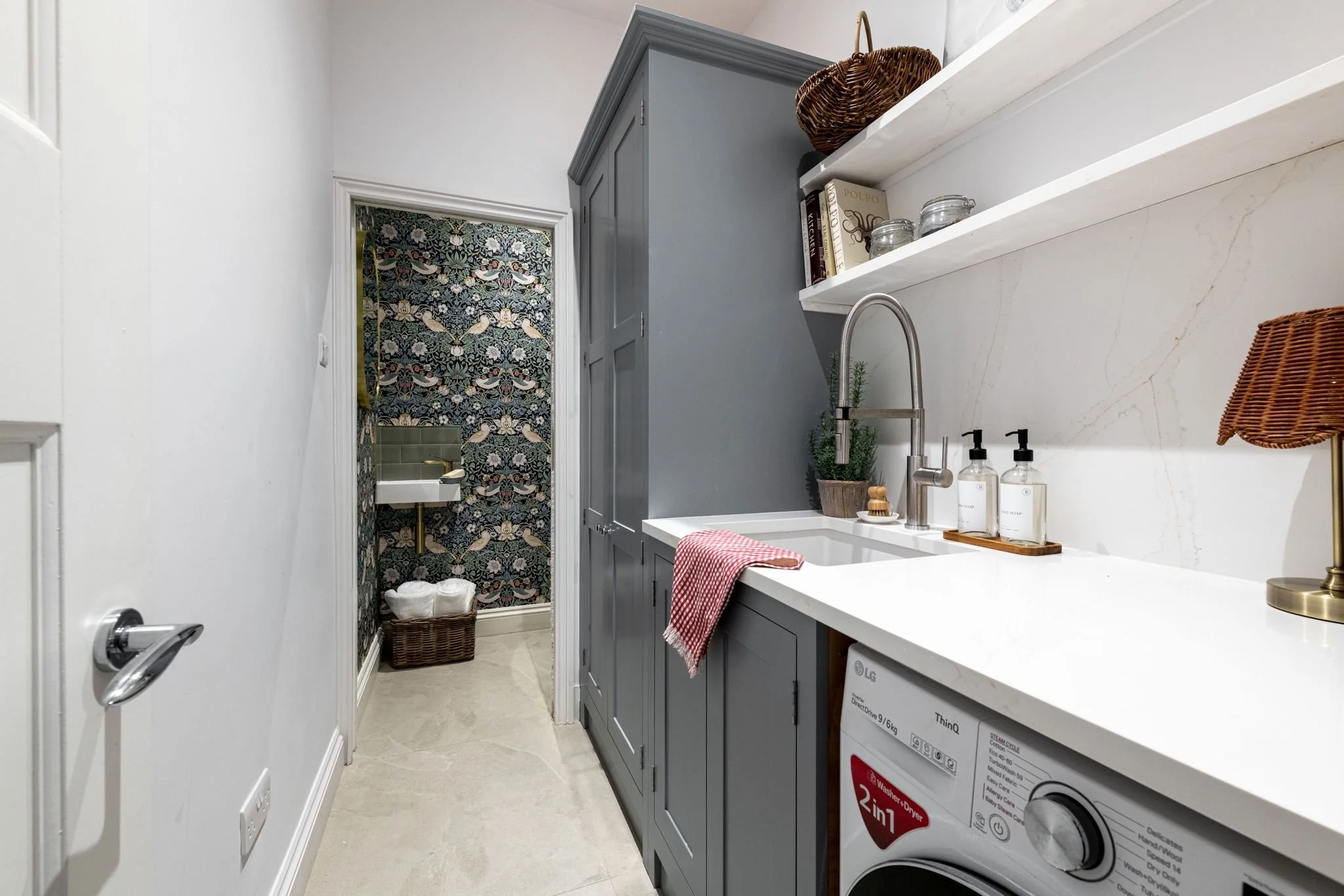 A small laundry area with a white countertop, gray cabinets, a laundry machine, and shelves with decorative items. At the back, there is a door with floral and bird wallpaper visible through the doorway.