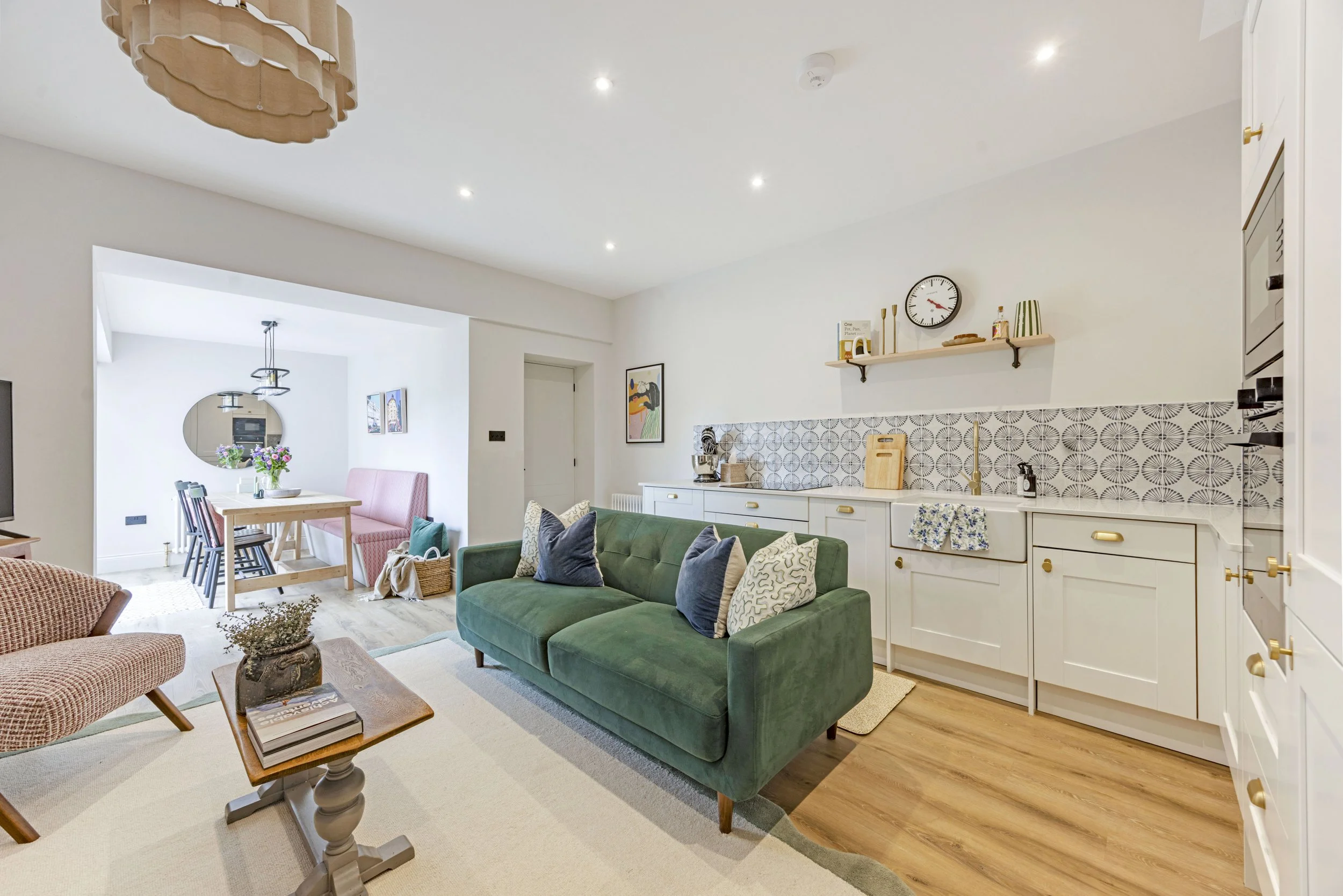 A living room and dining area with white walls, wooden flooring, a green sofa with patterned cushions, a wooden coffee table with books, a pink upholstered bench, a dining table with chairs, artwork on the walls, a wall clock, a shelf with decorative