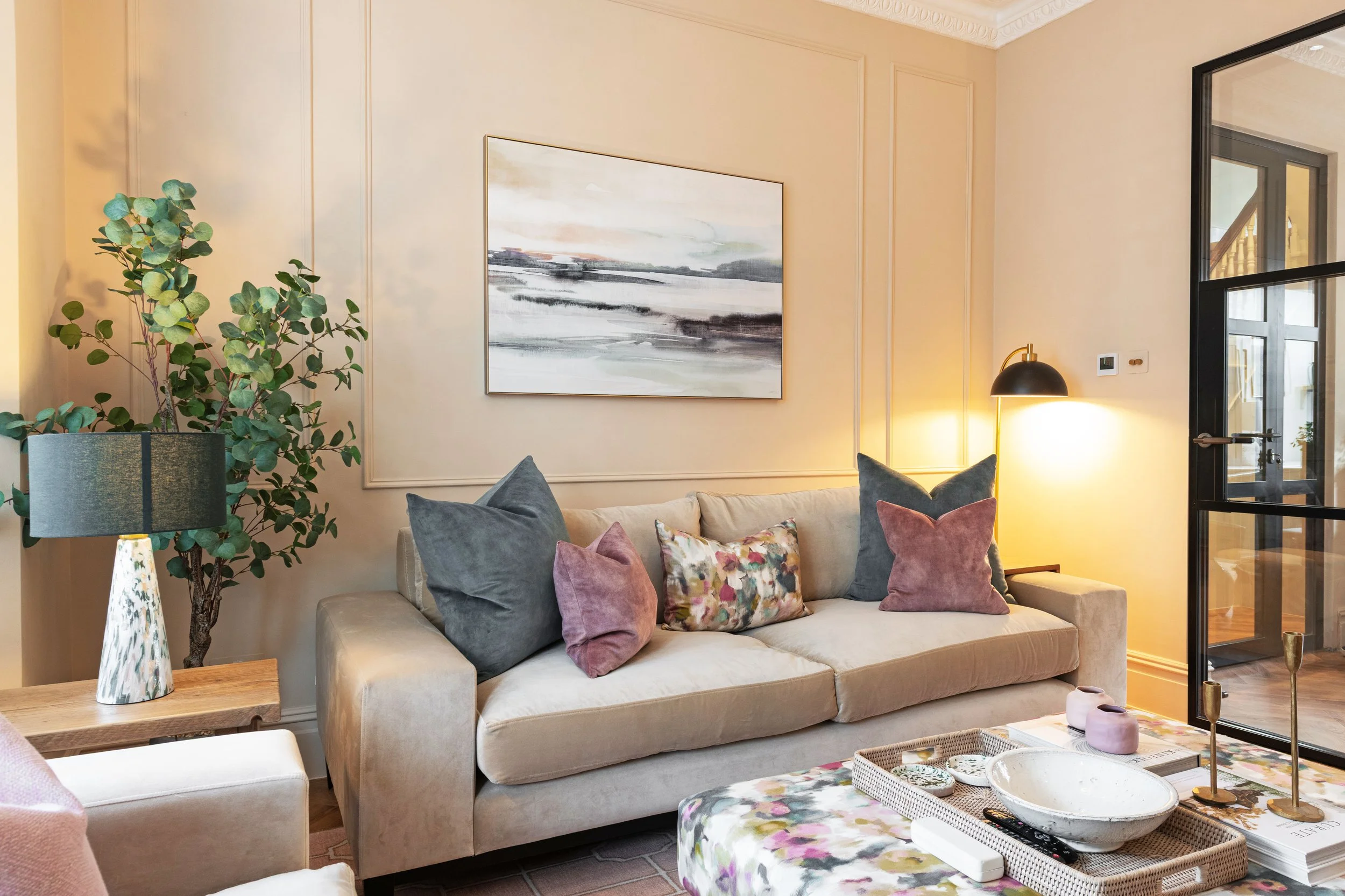 A living room with a beige sofa decorated with multi-colored pillows. There's a side table with a large potted green plant and a lamp. The wall behind the sofa has beige paneling and a large abstract landscape painting. To the right, there's a black-