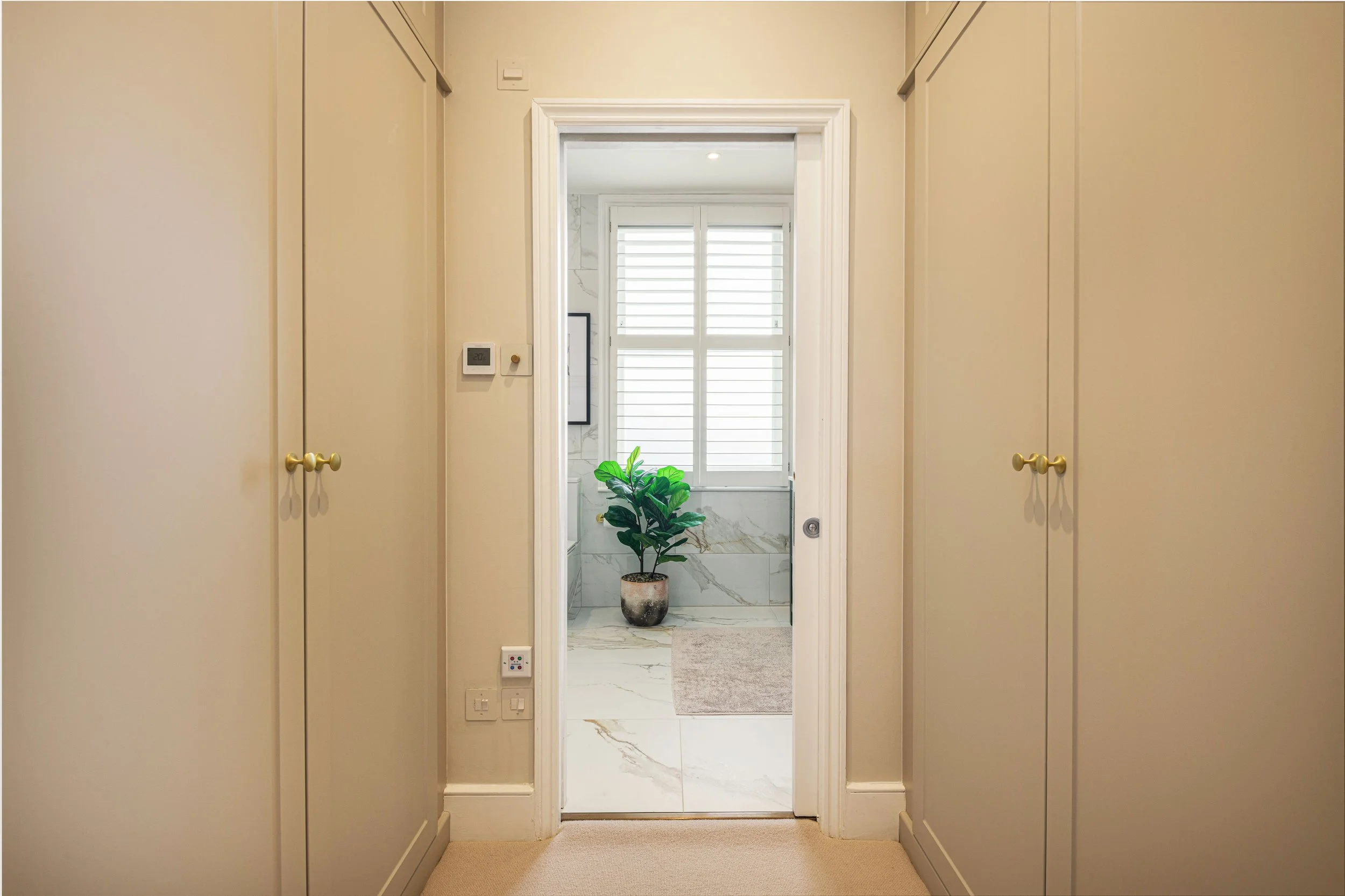 A hallway leading to a bathroom with a potted plant on the floor, white marble tiles, and white window shutters.
