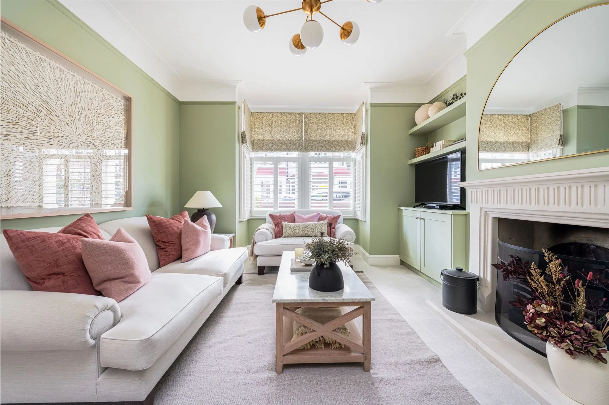 Living room with light green walls, a white couch with pink and beige pillows, a white armchair, a marble-topped coffee table with a black vase, a fireplace, a wall-mounted TV, shelves with decorative items, large windows with blinds, and a unique ce