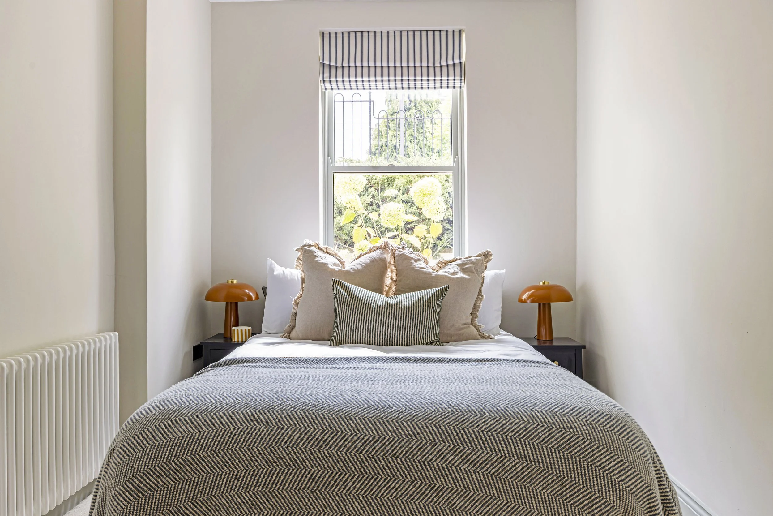 A neatly made bed with a black and white geometric patterned duvet, multiple pillows in beige, white, and striped covers, two orange bedside lamps, and a small black side table against a white wall with a tall window and striped valance showing green