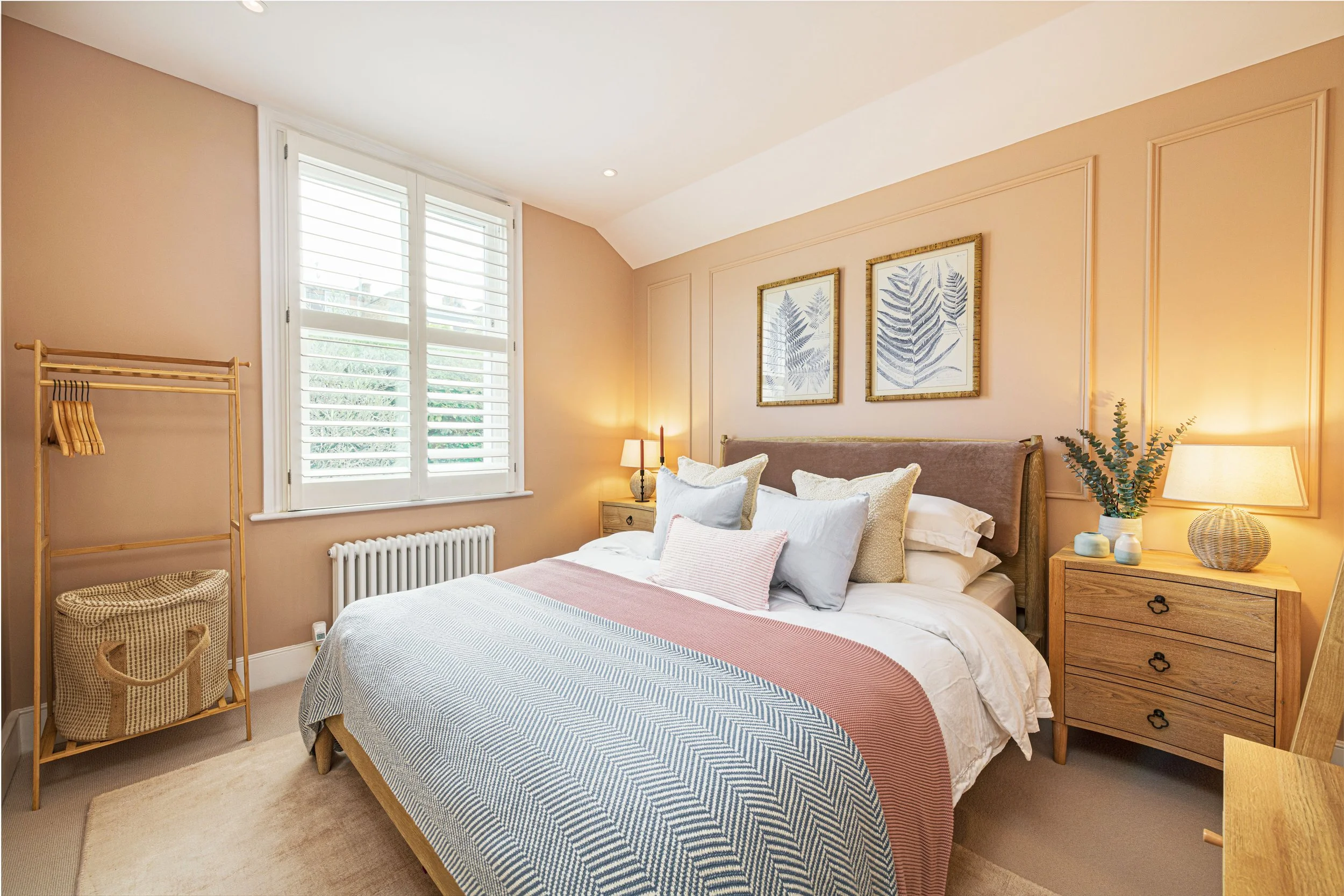 A cozy bedroom with a large bed with white and pastel-colored bedding, a wooden nightstand with a lamp, framed botanical prints on the wall, a window with white shutters, and a wooden clothing rack with baskets underneath.