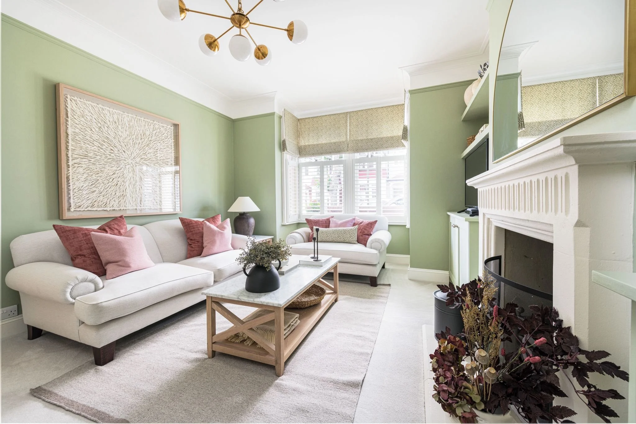 Living room with pastel green walls, white sofa with pink cushions, wooden coffee table, and fireplace. Large window with white blinds and decorative window treatments. Modern chandelier hanging from the ceiling.