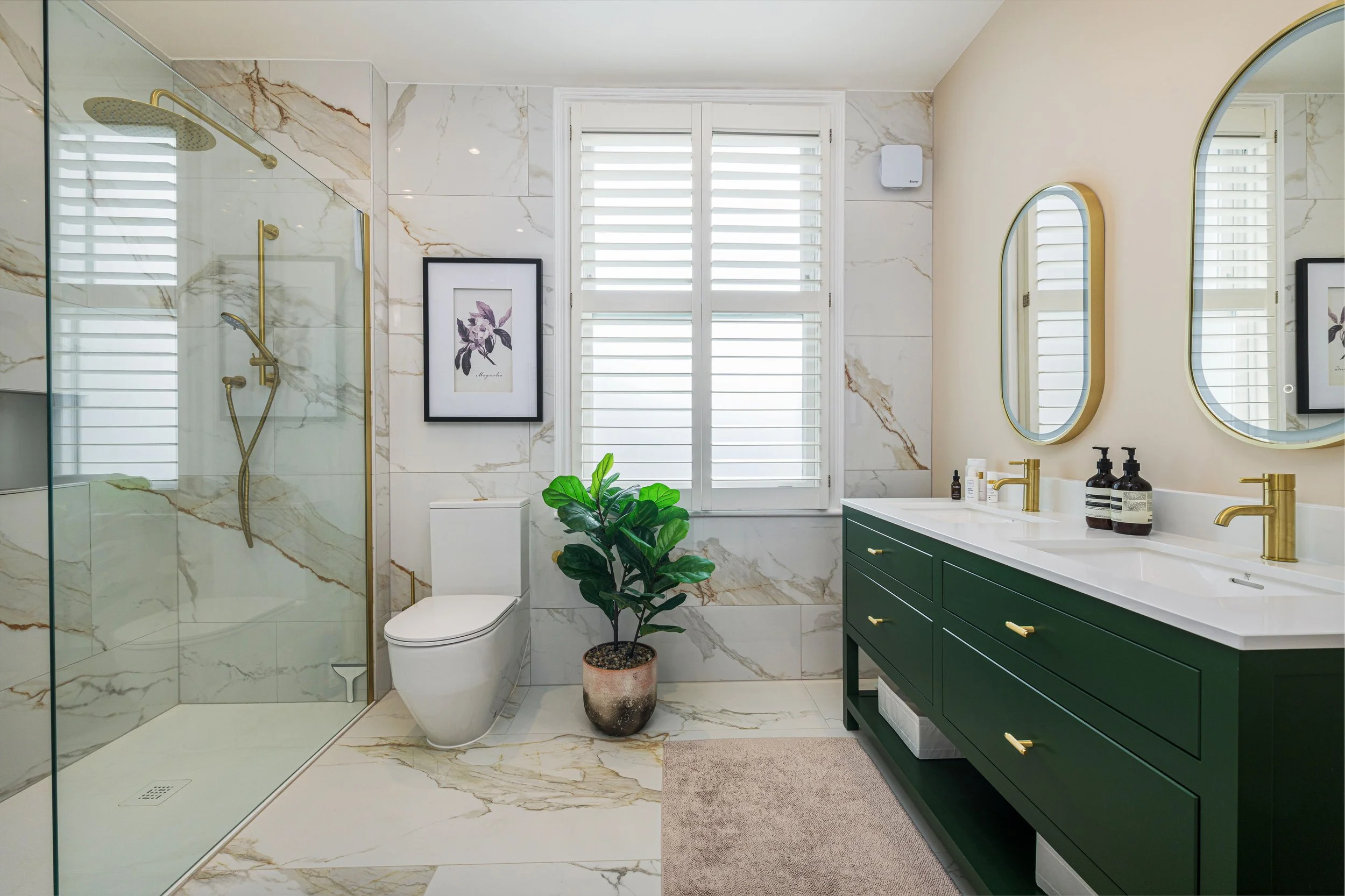 Modern bathroom featuring a glass-enclosed shower with gold fixtures, marble walls and floors, two round mirrors above a dark green vanity, a plant in a pot, and artwork on the wall.