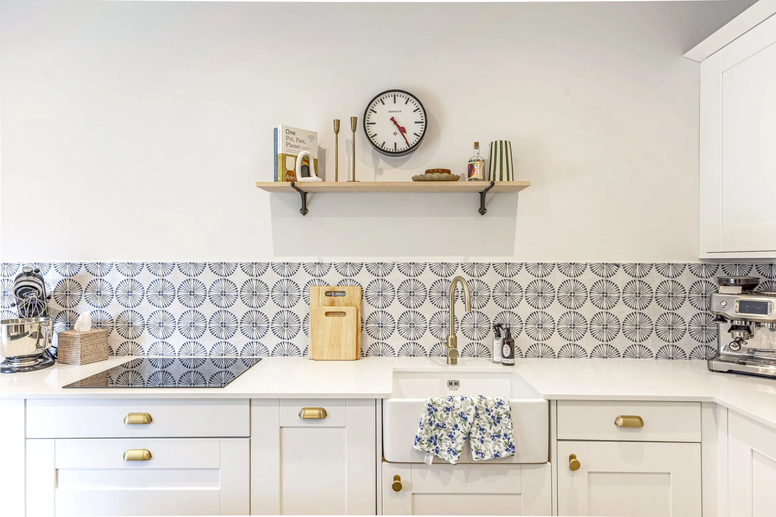 Kitchen with white cabinets, a white sink with a floral dish towel, a wooden cutting board, a gold faucet, a patterned blue and white backsplash, a floating wooden shelf with books, decorations, and a clock, a stand mixer, and a toaster.