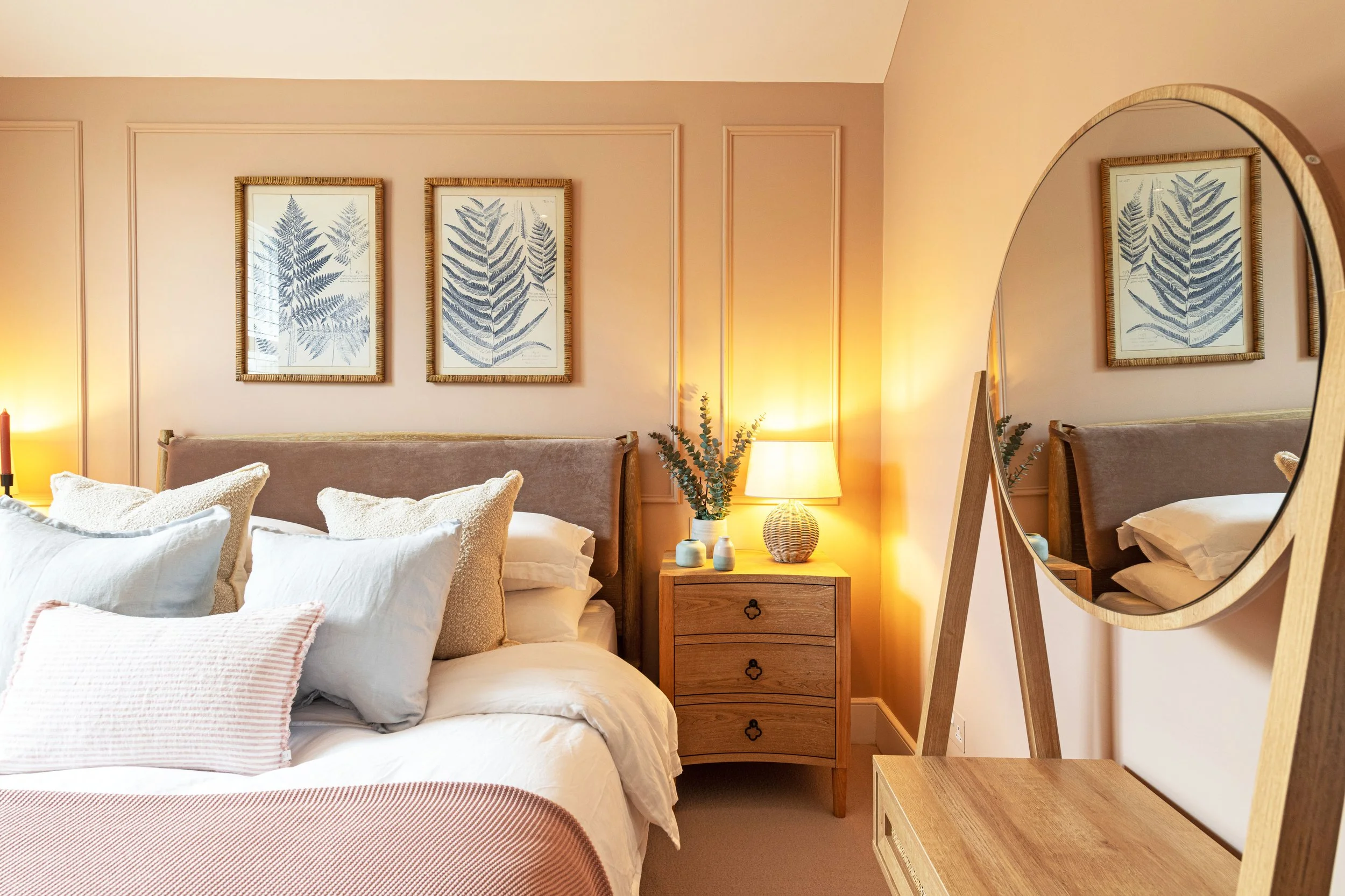 A cozy bedroom with a bed adorned with white and pink pillows, a wooden nightstand with decorative vases, a lamp, framed botanical prints on the wall, a large circular mirror reflecting the room, and soft warm lighting.
