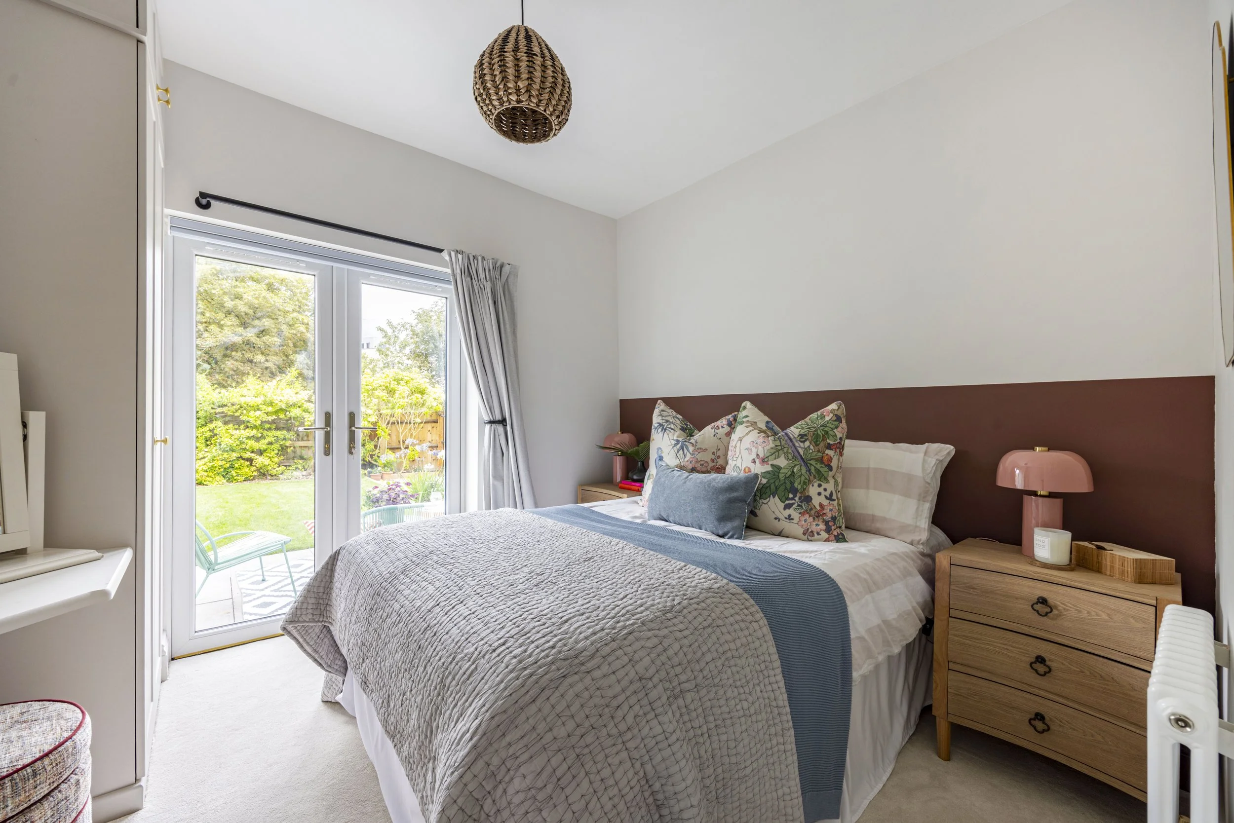A cozy bedroom with a bed, floral pillows, a bedside table with a pink lamp, and a large window with white curtains showing a garden outside.