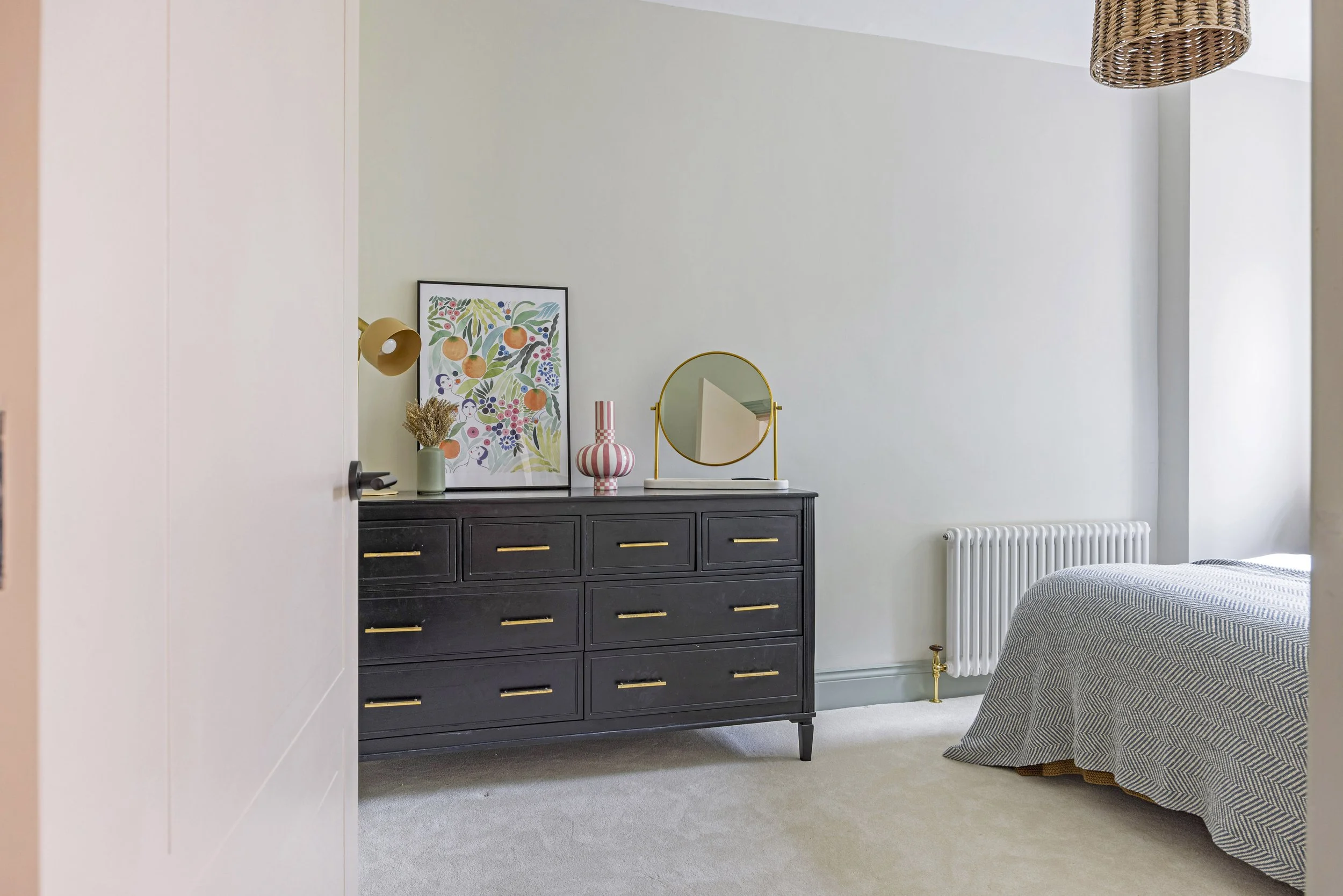 A bedroom with a black dresser, decorative items including a framed botanical print, a gold wall-mounted lamp, a striped pink and white vase, a round gold mirror, a white radiator, and part of a bed with a gray and white striped blanket. The room has