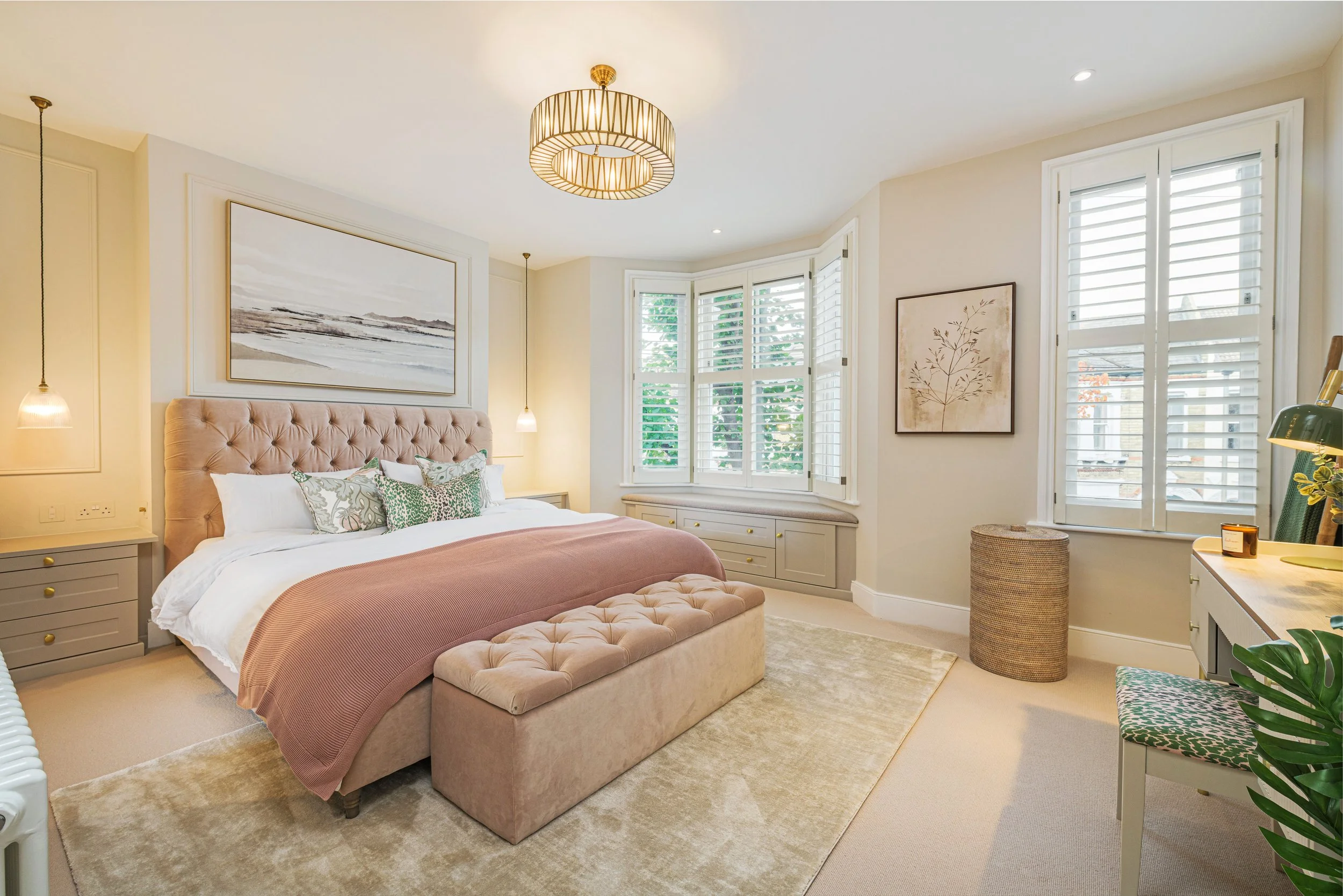 A bedroom with a large bed, tufted headboard, and pillows. There are large windows with shutters and wall art. A cream rug covers the floor, and there are side tables and a bench at the foot of the bed.