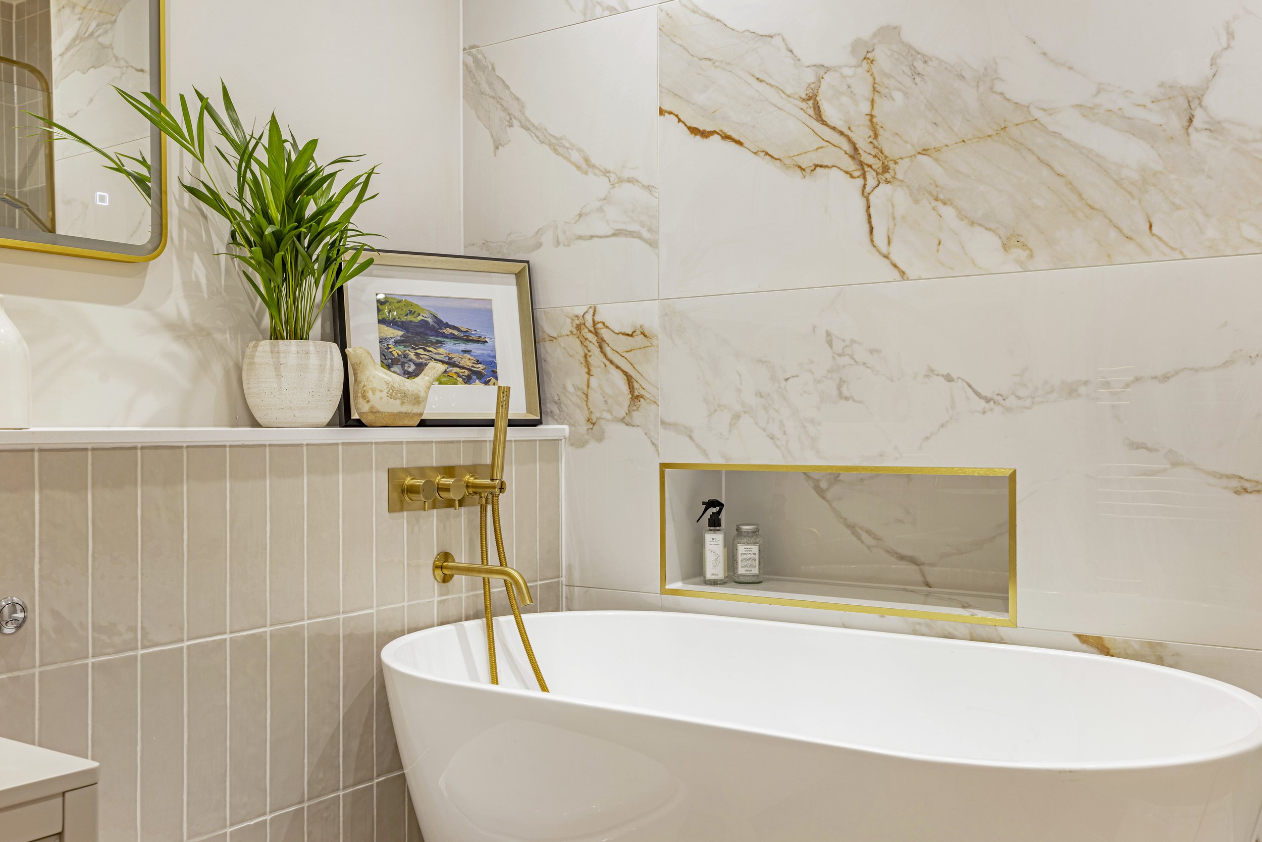 A modern bathroom with a white freestanding bathtub, gold fixtures, a large green plant in a beige pot, a framed landscape painting, a mirrored wall, and marble tiles.