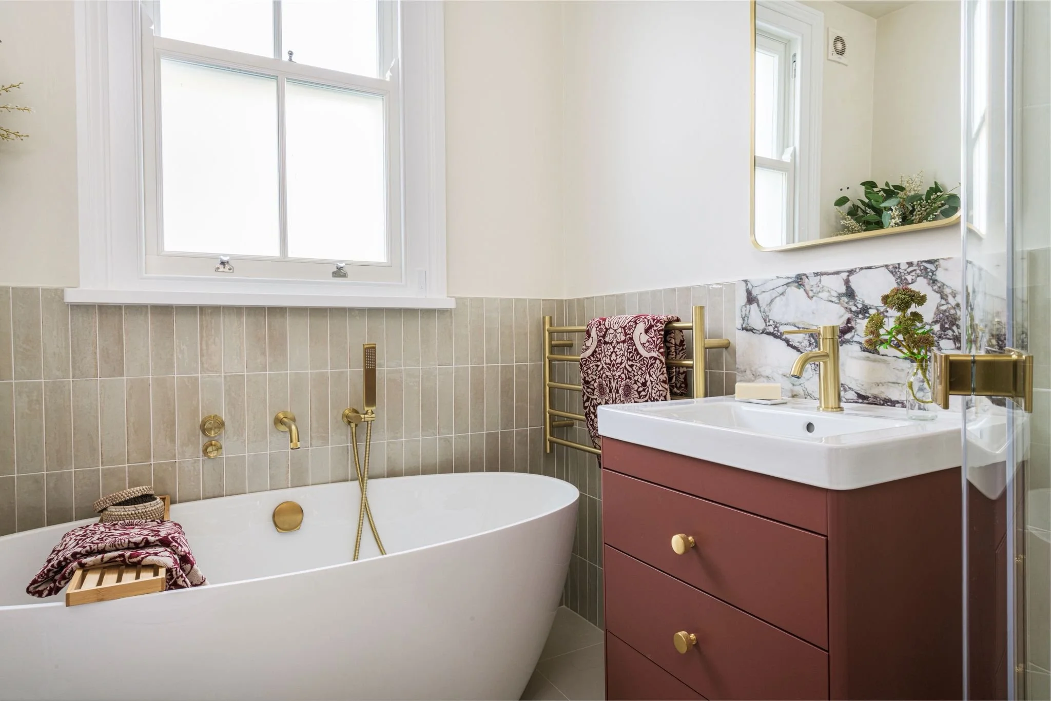 A bathroom with a white bathtub, beige tile walls, a large window, a white sink with a pink cabinet, gold fixtures, a towel rack with a patterned towel, a mirror, and some plants.