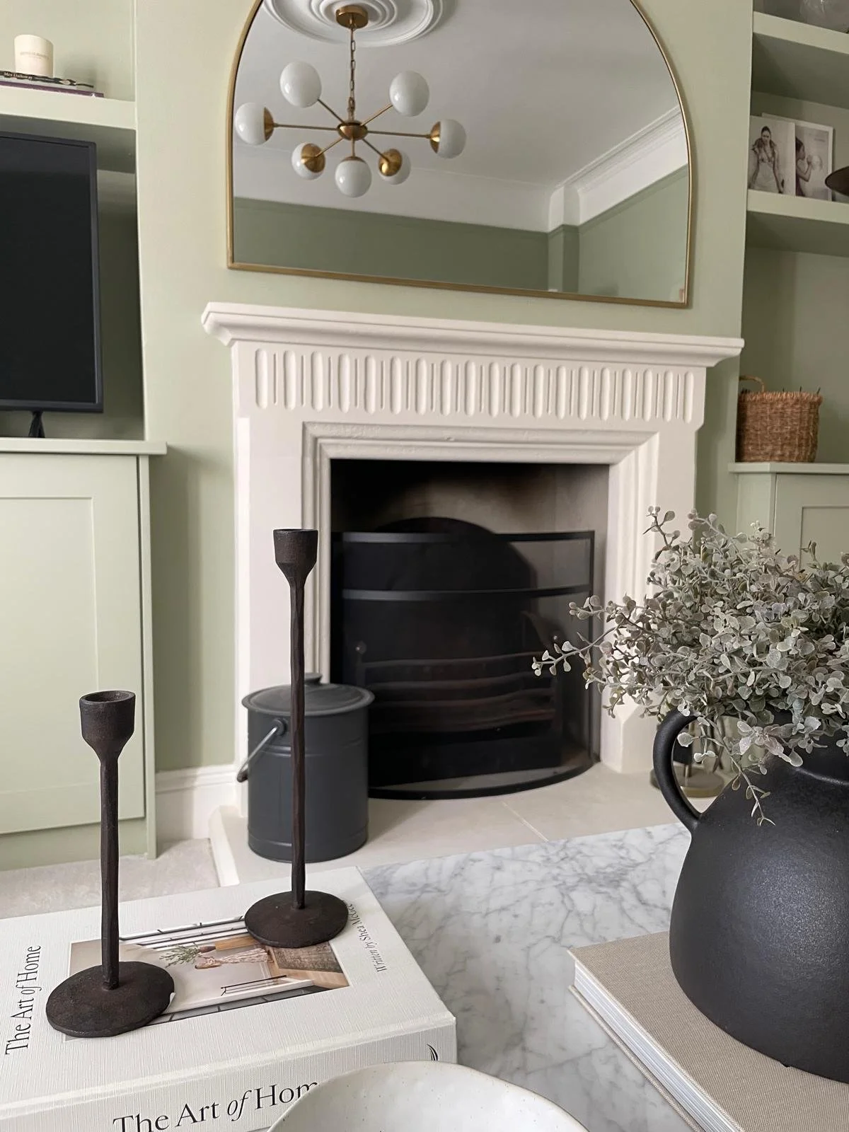 Decorative living room with a white fireplace, a large mirror above it, and a modern brass chandelier. In front of the fireplace, there are two black candle holders, a black vase with dried flowers, and a gray bucket. A white book titled 'The Art of 