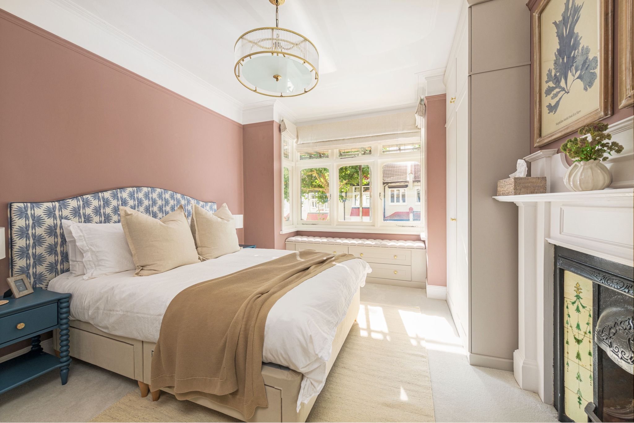A cozy bedroom with a large bed that has a blue patterned headboard, beige pillows, and a beige blanket. There is a blue nightstand with picture frames and a clock next to the bed. The room has a big window with a view of trees and neighboring houses