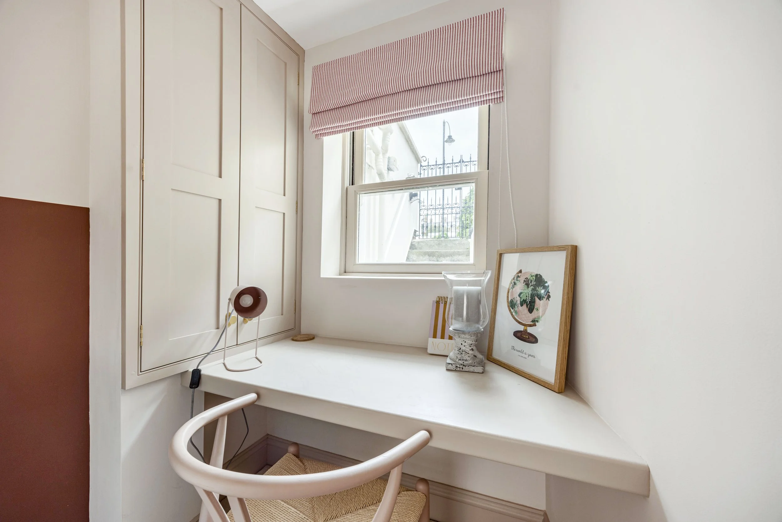 A small workspace corner with a white built-in desk by a window with a red and white striped Roman shade. There is a framed botanical print, a glass candle holder, and a framed art piece on the desk. A white chair with a woven seat is pulled up to th