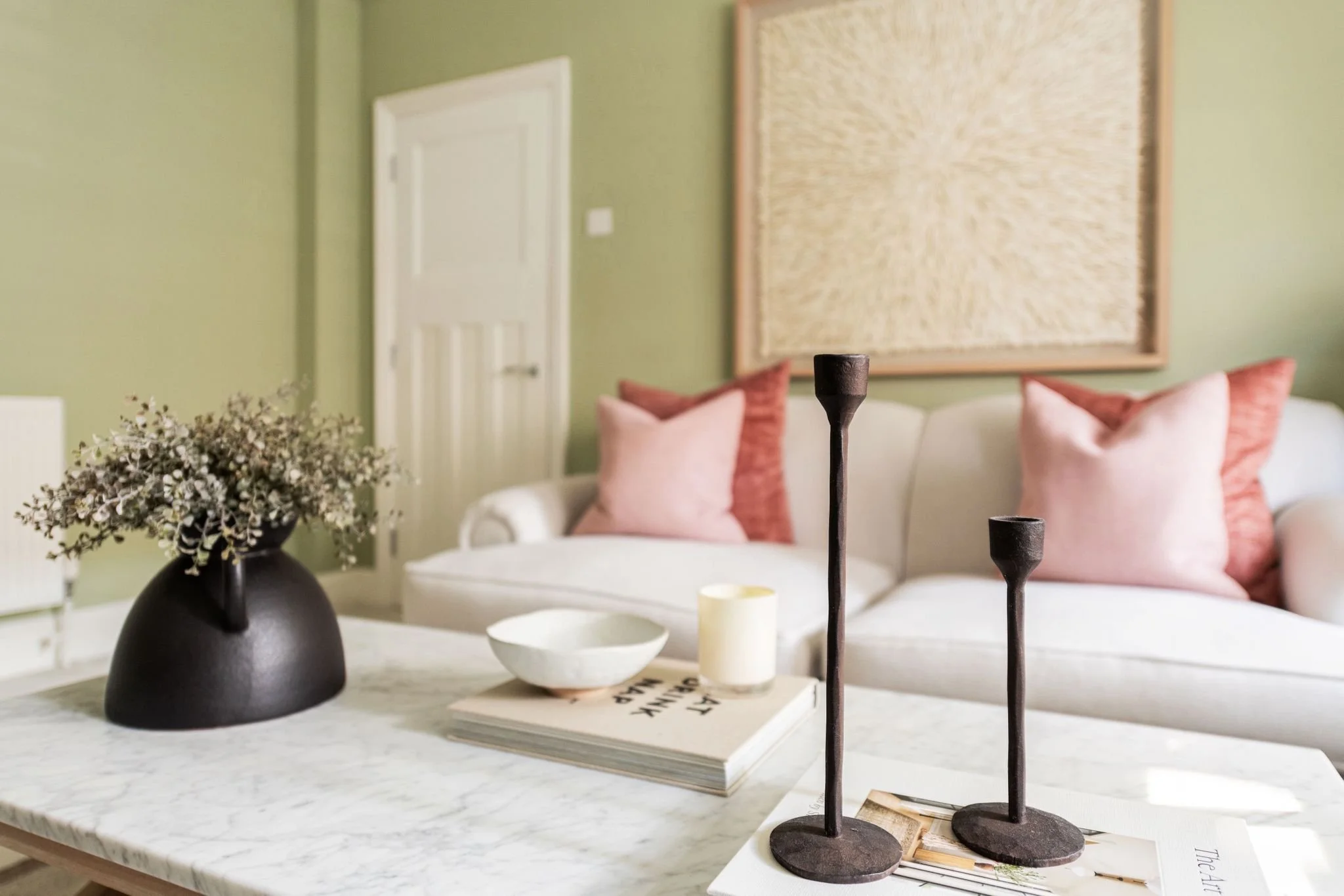 Living room with green walls, white door, abstract art, pink throw pillows on white sofa, black vase with dried flowers, and black candle holders on a white marble table.