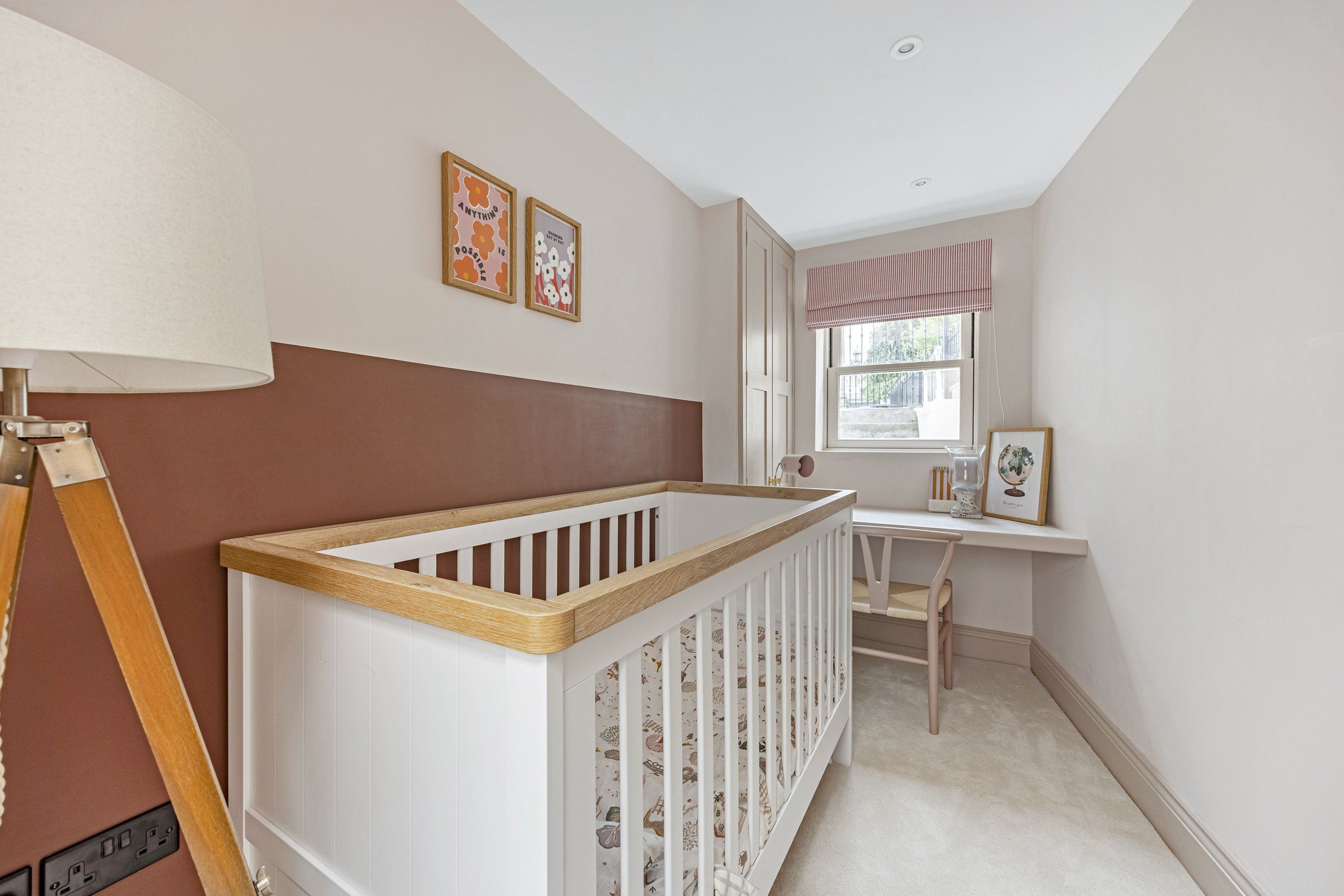 Nursery room with a white crib, framed pictures on the wall, a window with red striped valance, and a desk with a globe and chair.