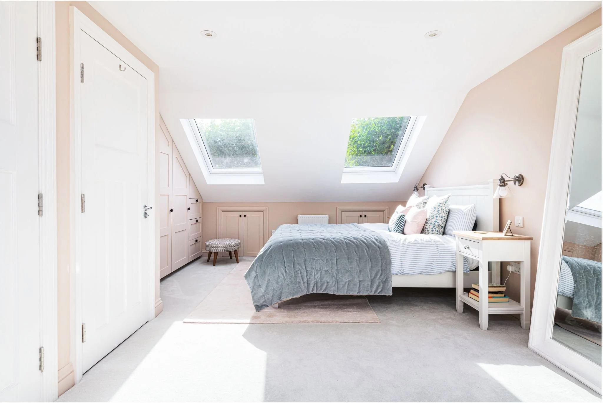 A cozy bedroom with sloped ceilings and two skylight windows showing greenery outside. The bed has a white headboard and is made with a gray quilt, surrounded by decorative pillows. There is a small white nightstand with books and a mirror nearby, an