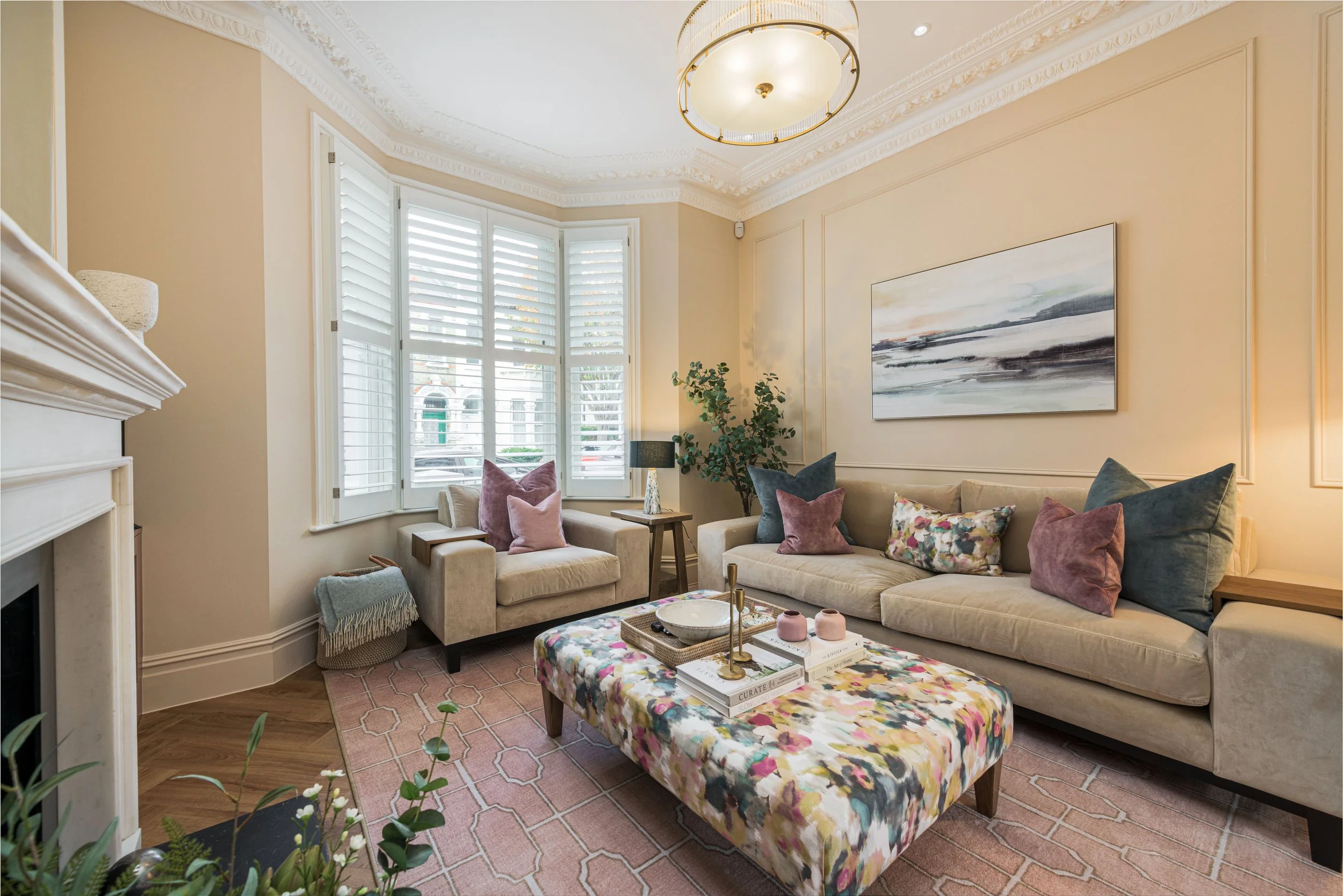 Living room with large beige sofas, colorful pillows, a floral accent chair, a wooden coffee table with decorative items, a pink patterned rug, a large window with white shutters, and an abstract painting on the wall. There is a green potted plant an