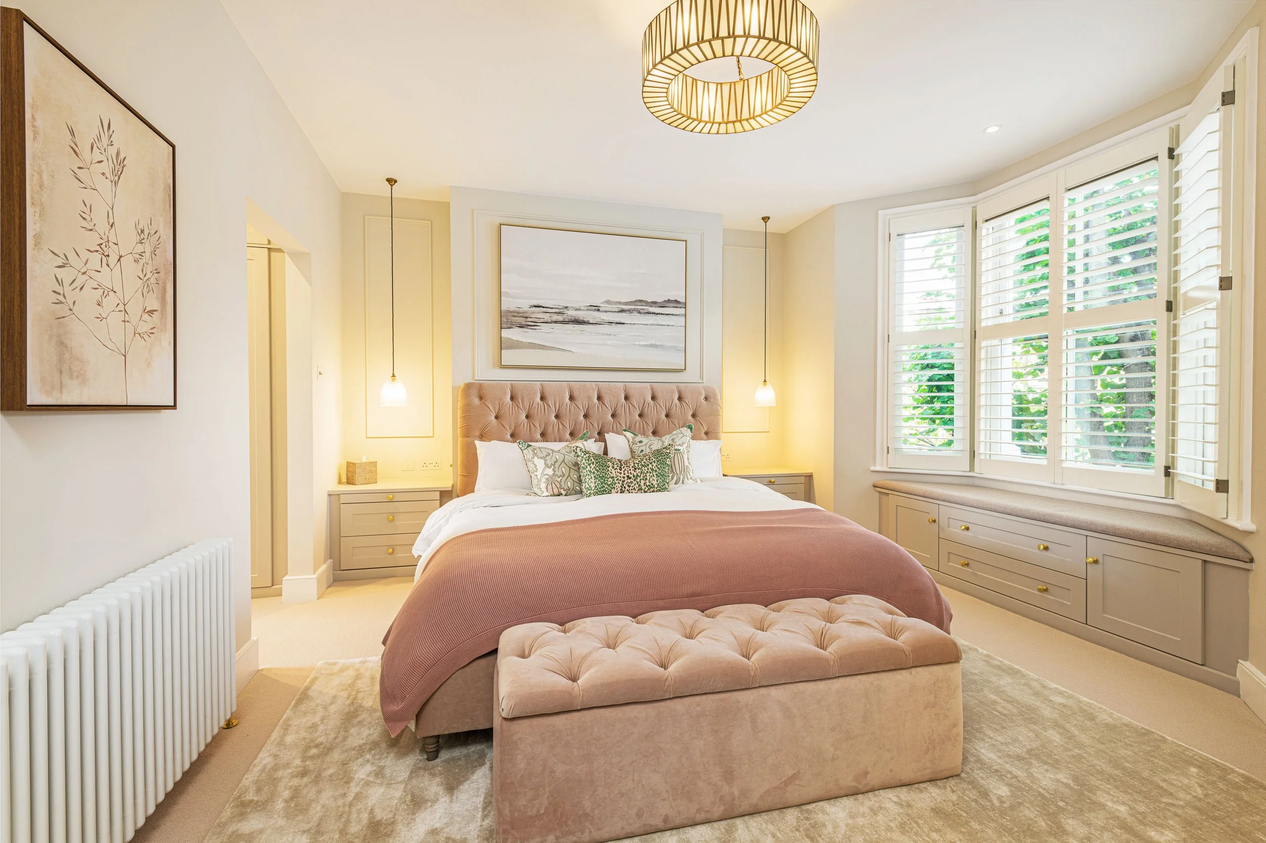 A bedroom with a bed, at the foot of the bed is a pink tufted bench. The bed has a tufted pink headboard and is made with white sheets, decorative pillows, and a mauve bedspread. There are two nightstands with lamps, framed beach artwork above the be