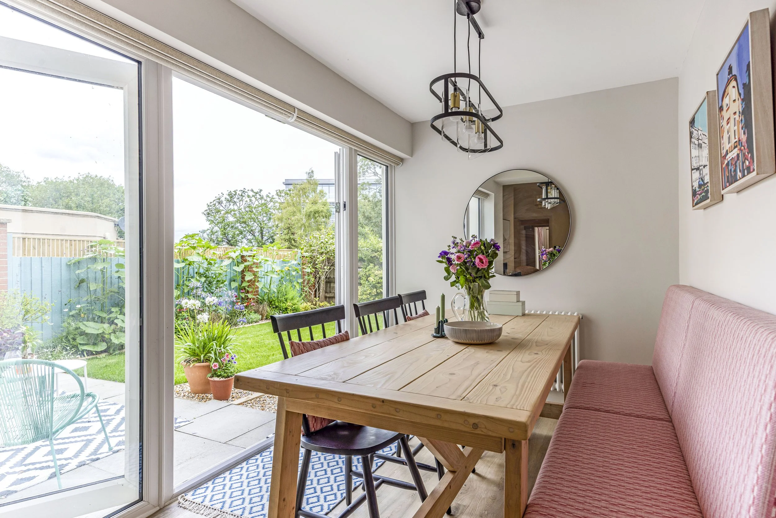 A dining area with a wooden table and a pink upholstered bench. A bouquet of pink and purple flowers is on the table. Large glass sliding doors open to a garden with potted plants and a patio. Inside, a round mirror and framed artwork are on the whit