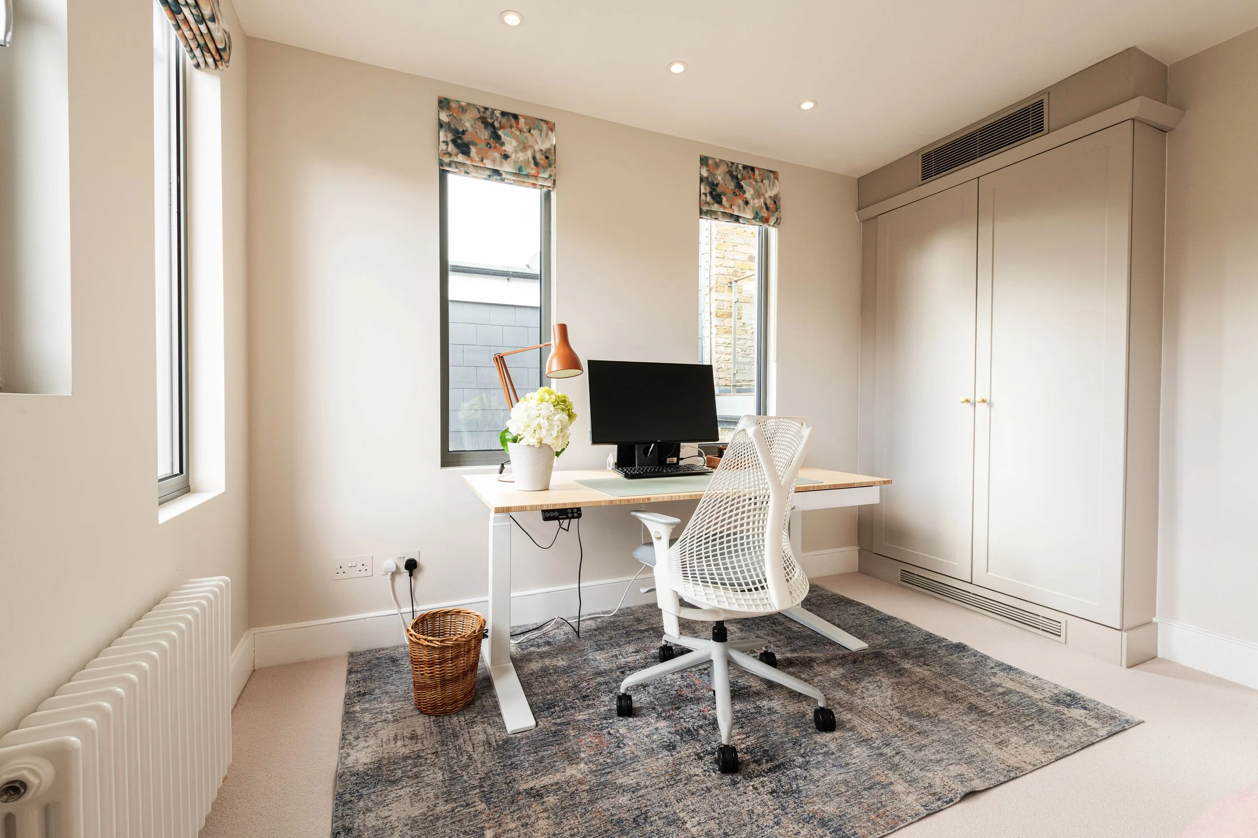 Home office with a desk, computer, white office chair, flower vase, and a desk lamp near a window. The room has beige walls, a gray and blue rug, built-in closet, and small window roman blinds.
