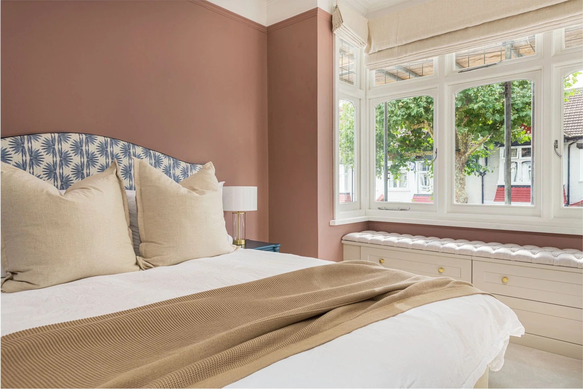 A bedroom with a bed featuring beige pillows, a tan bed runner, and a patterned headboard. There is a window seat below a large window with white frames and cream roman shades, revealing green trees outside.