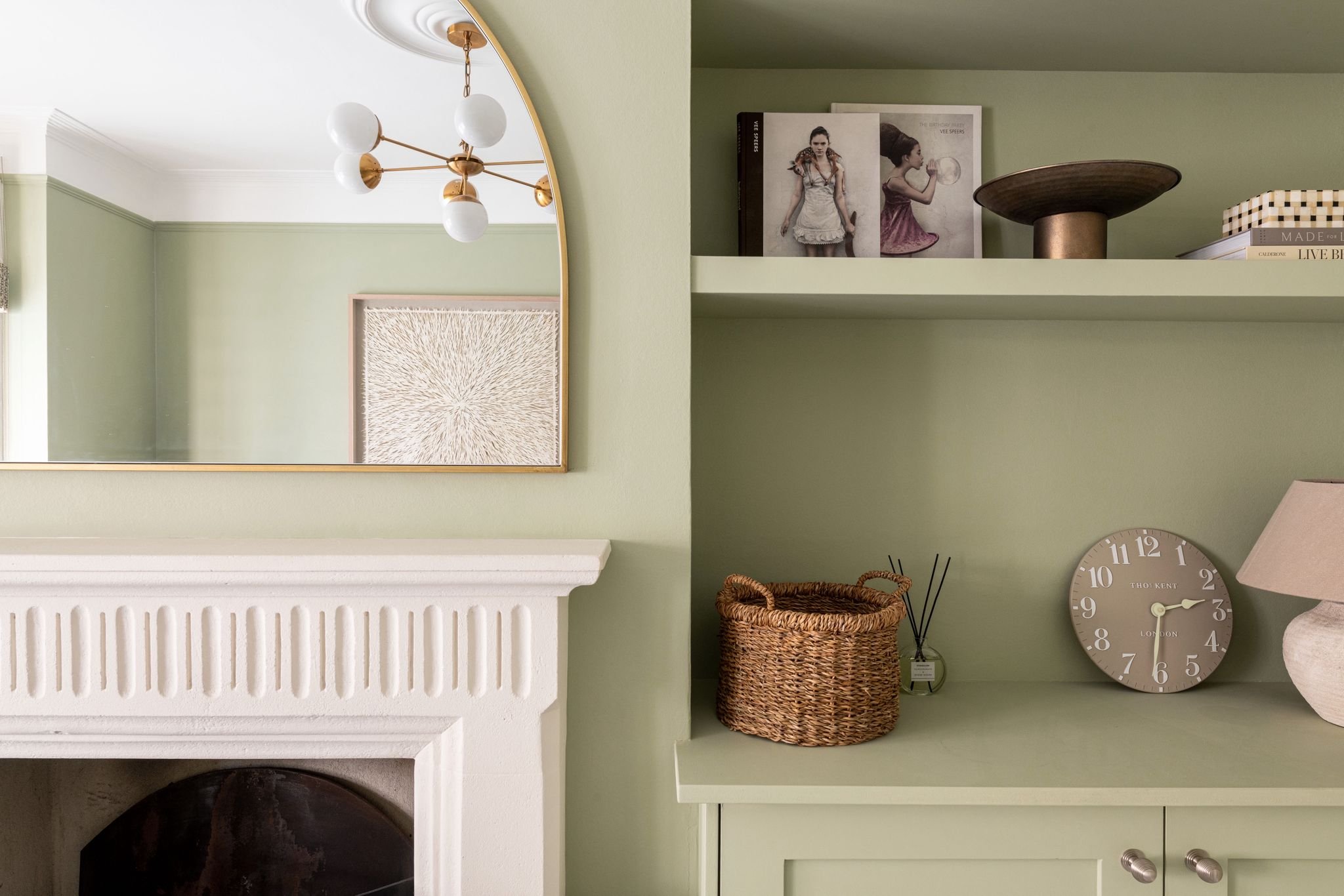 A living room corner with a large round mirror, a white fireplace, a green wall with wall shelves holding books and decor, a woven basket, a clock, and a table lamp.