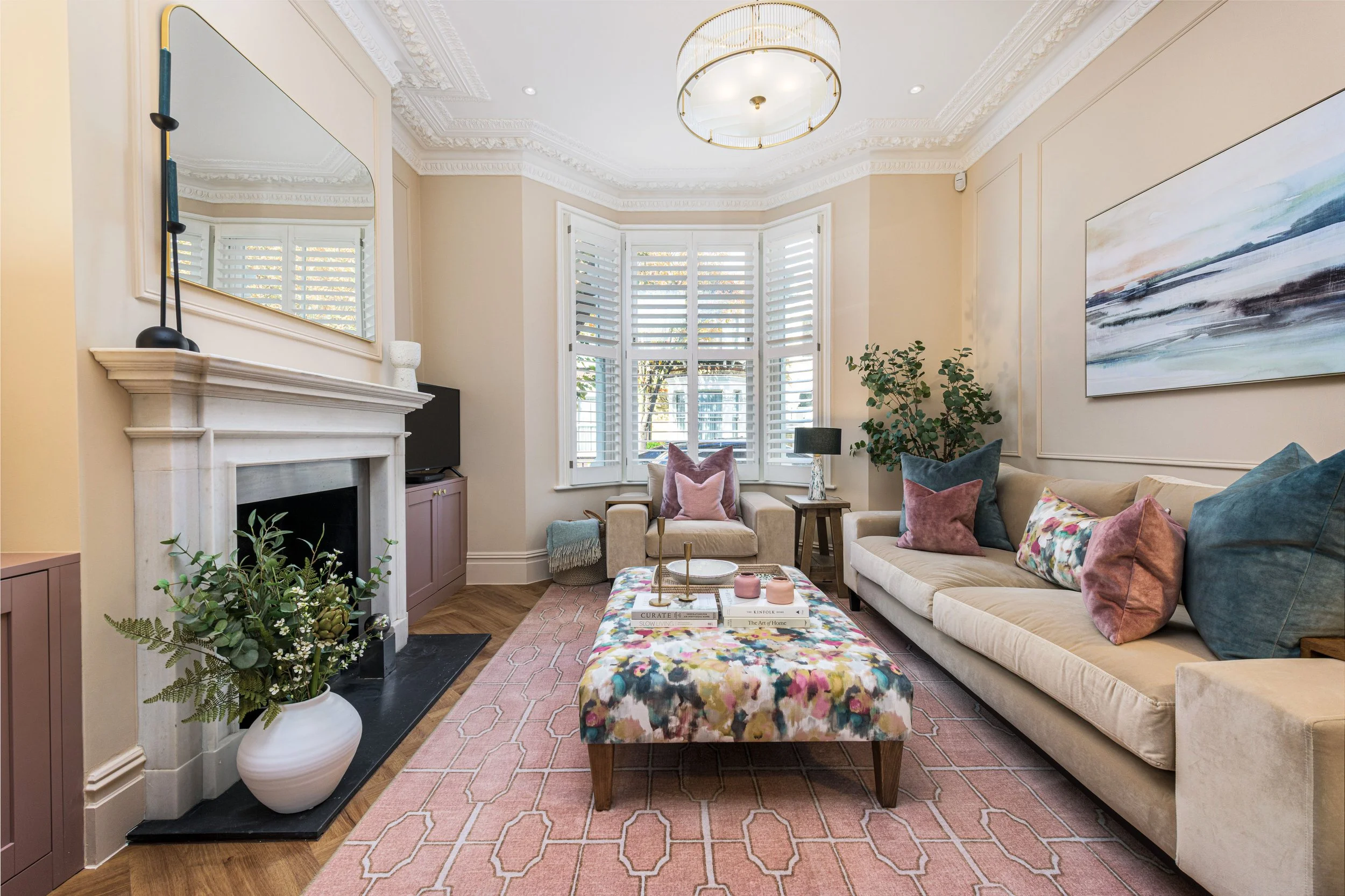 A living room with a beige sectional sofa, colorful pillows, a floral upholstered ottoman, a pink patterned rug, a stone fireplace with a large mirror, plants, wall art, and sunlight through white plantation shutters.