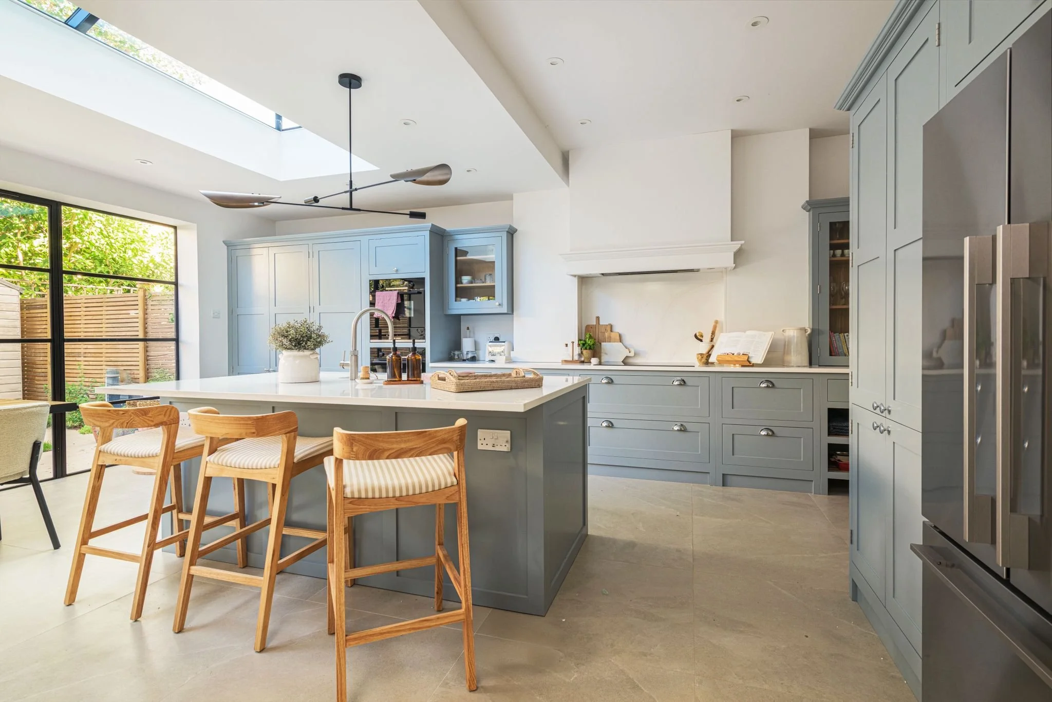 Modern kitchen with light blue cabinets, white countertops, wooden chairs around a kitchen island, large windows with outdoor view, and a skylight ceiling.