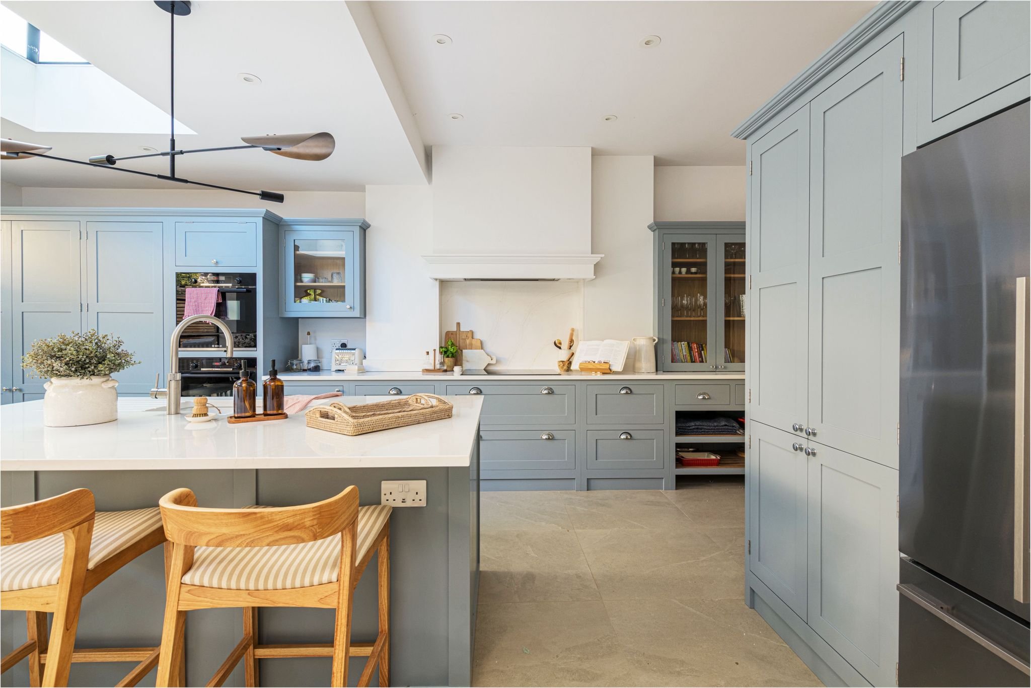 Modern kitchen with blue cabinets, an island with a white countertop, wooden chairs, and various kitchen items including a plant, bottles, and a woven tray.