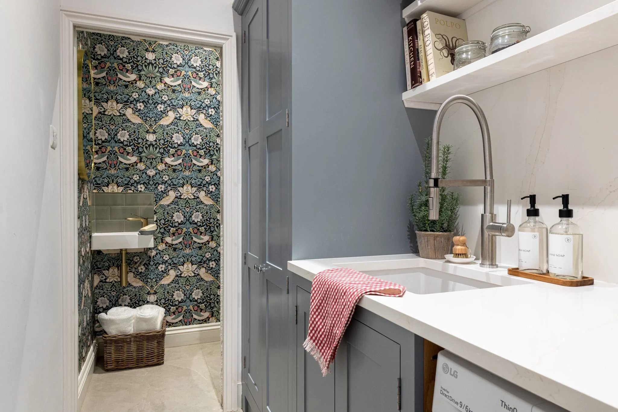 A bathroom with a decorative wallpapered shower area featuring birds and flowers, a small white sink with a brass faucet, and a wicker basket on the floor. To the right, a gray cabinet with a white countertop holds soap dispensers, a potted plant, an