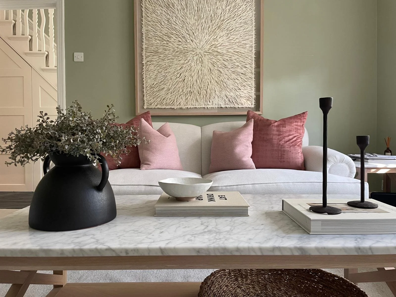 Living room with a beige sofa adorned with pink and rust-colored pillows, a large abstract wall hanging, a black vase with greenery, a bowl and magazines on a marble coffee table, and two black candle holders.