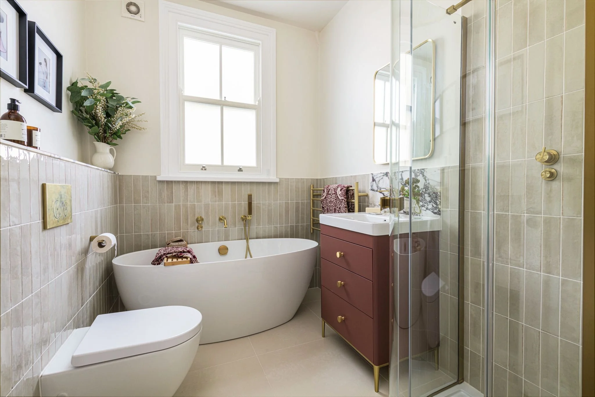 A bathroom with a large window, a freestanding bathtub, a vanity with a sink, and a glass shower enclosure. Decor includes framed pictures, a potted plant, a towel, and golden fixtures.