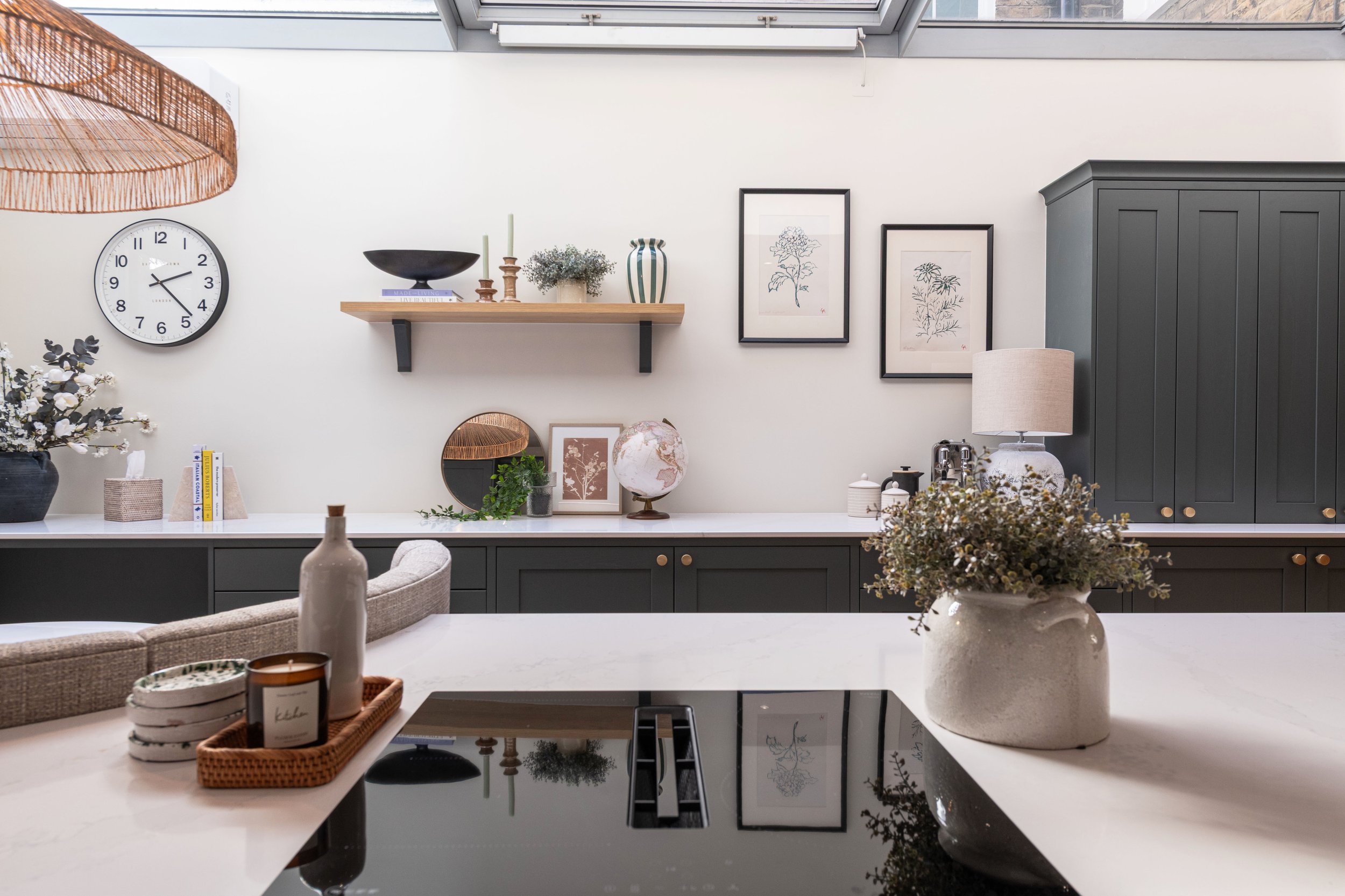 Interior of a modern kitchen with black cabinetry, framed botanical artwork, a white countertop with decorative items, and a wall shelf with vases and books.