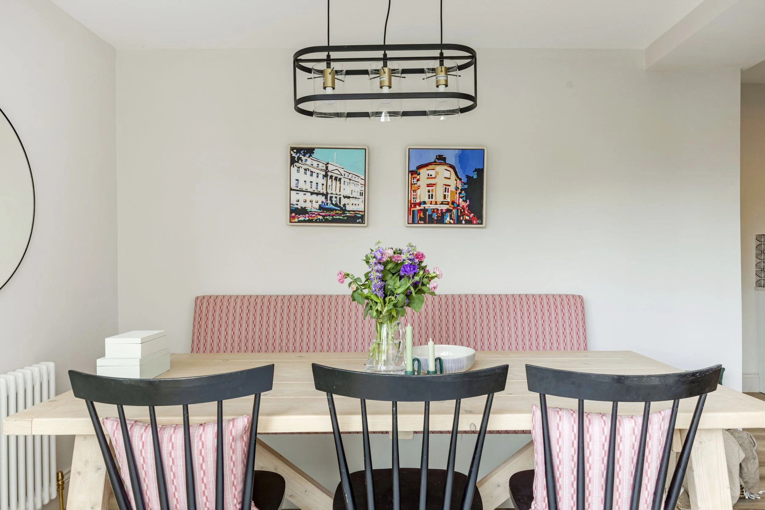 A dining room with a light wooden table, black chairs, a pink and white striped bench, a vase of pink, purple, and green flowers, two framed paintings of buildings on the wall, a modern black chandelier with glass and gold accents, and a white box on