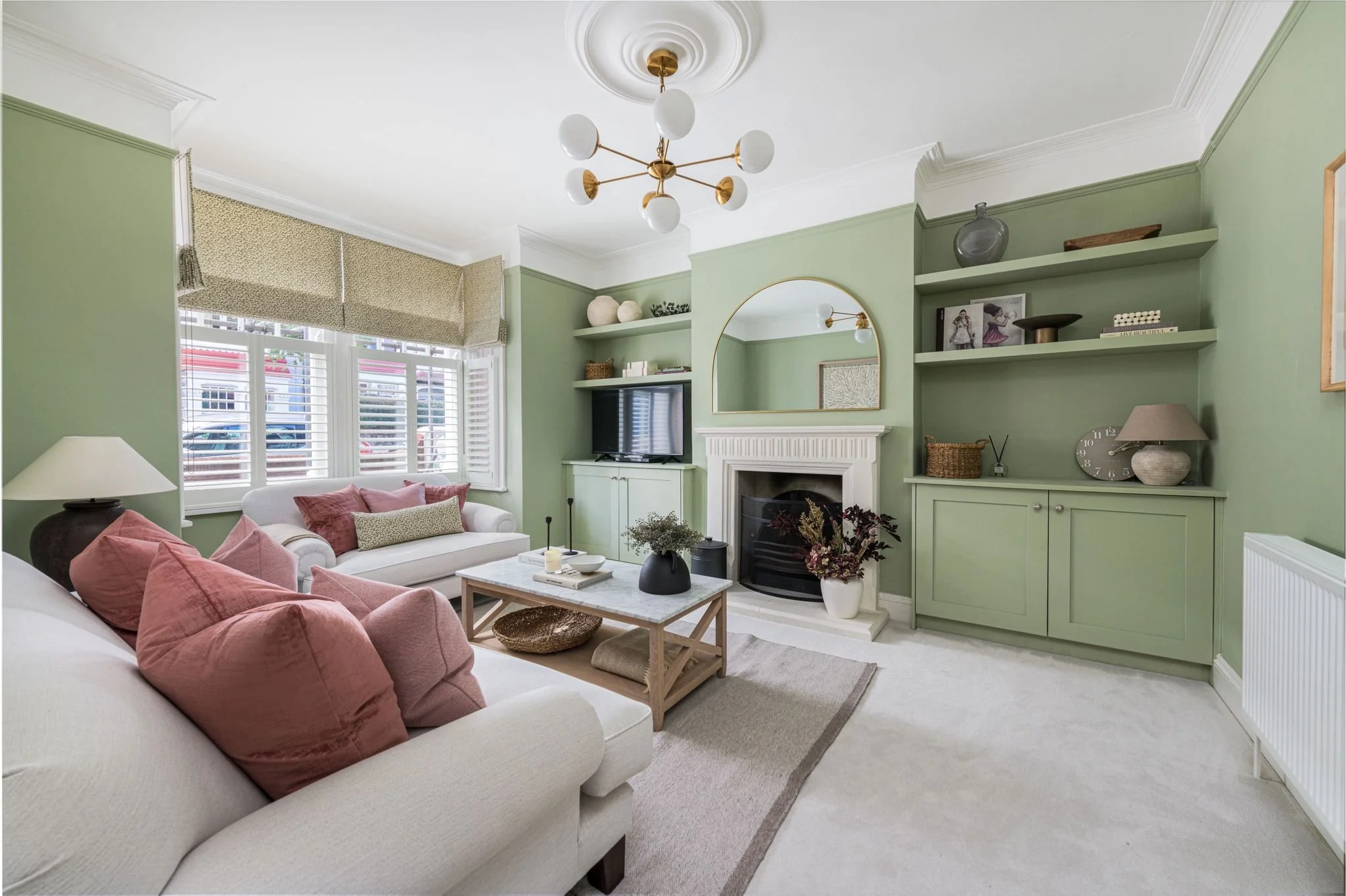 A bright living room with a white sofa, pink cushions, and a wooden coffee table. A fireplace with a gold-framed mirror above it, green built-in shelves with decorations, and a wall-mounted TV. A large window with blinds and Roman shades lets in natu