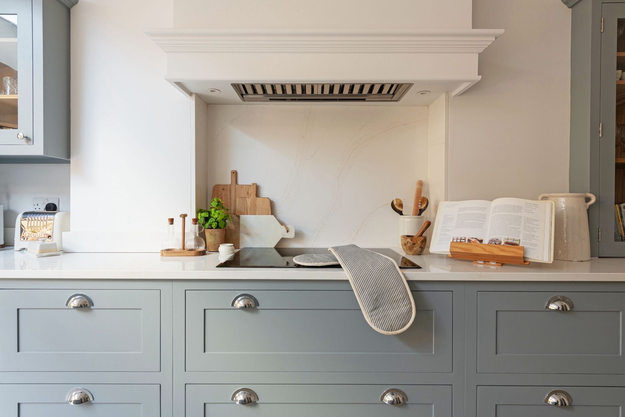 A kitchen countertop with a cookbook, wooden utensils, a potted plant, cutting boards, and a striped cloth, with a stove and cabinets in the background.