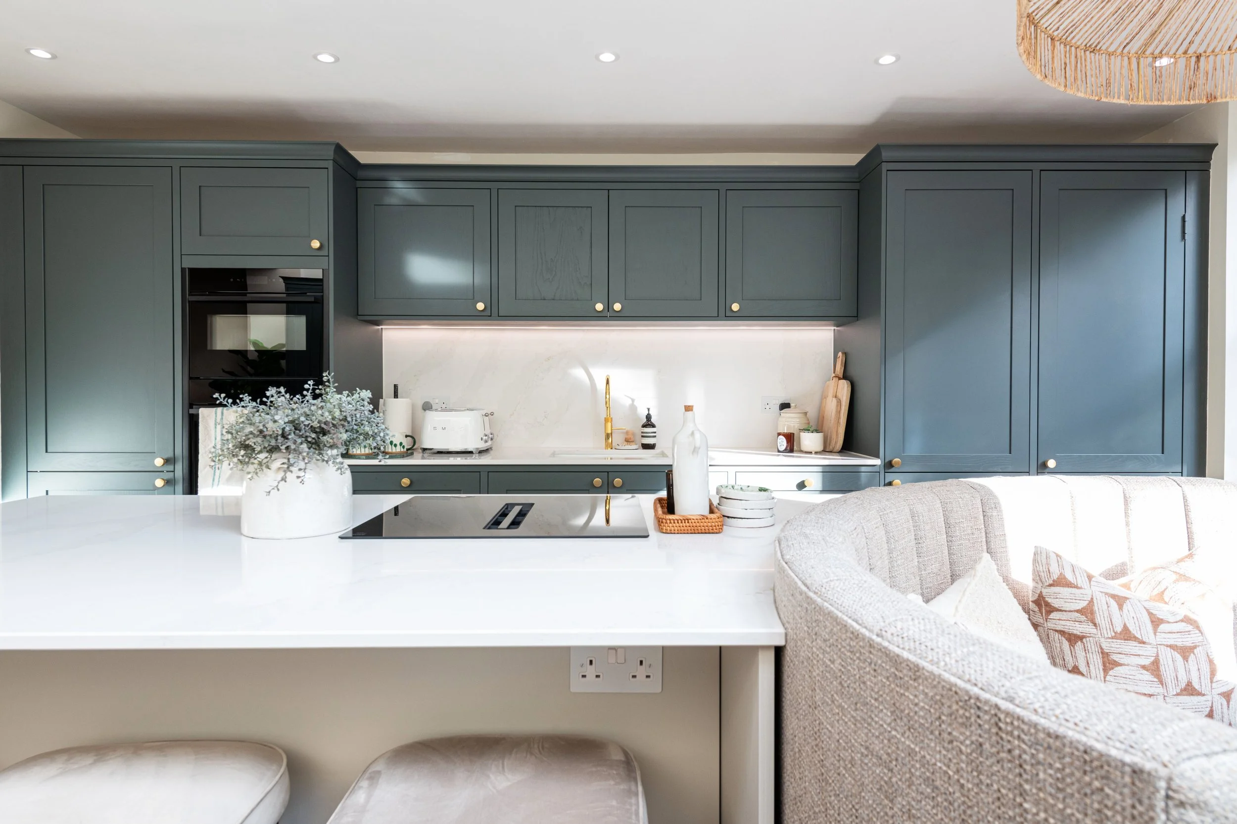 Modern kitchen with dark gray cabinets, white marble backsplash, and white countertop. Kitchen island has a potted plant, paper towel holder, and bottles. Part of a beige sofa with a patterned pillow is visible in the foreground.