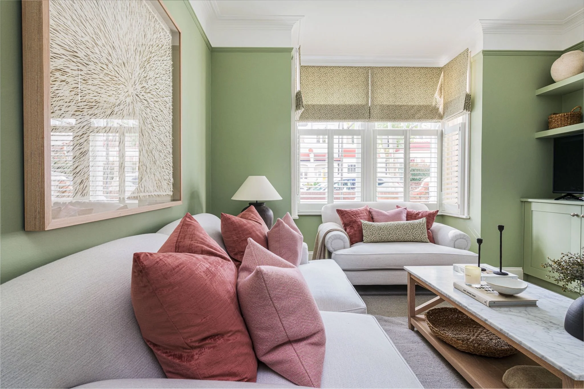 A cozy living room with light green walls, a white sofa, pink throw pillows, a coffee table with books and decor, a window with shutters and patterned valances, and a framed abstract artwork on the wall.