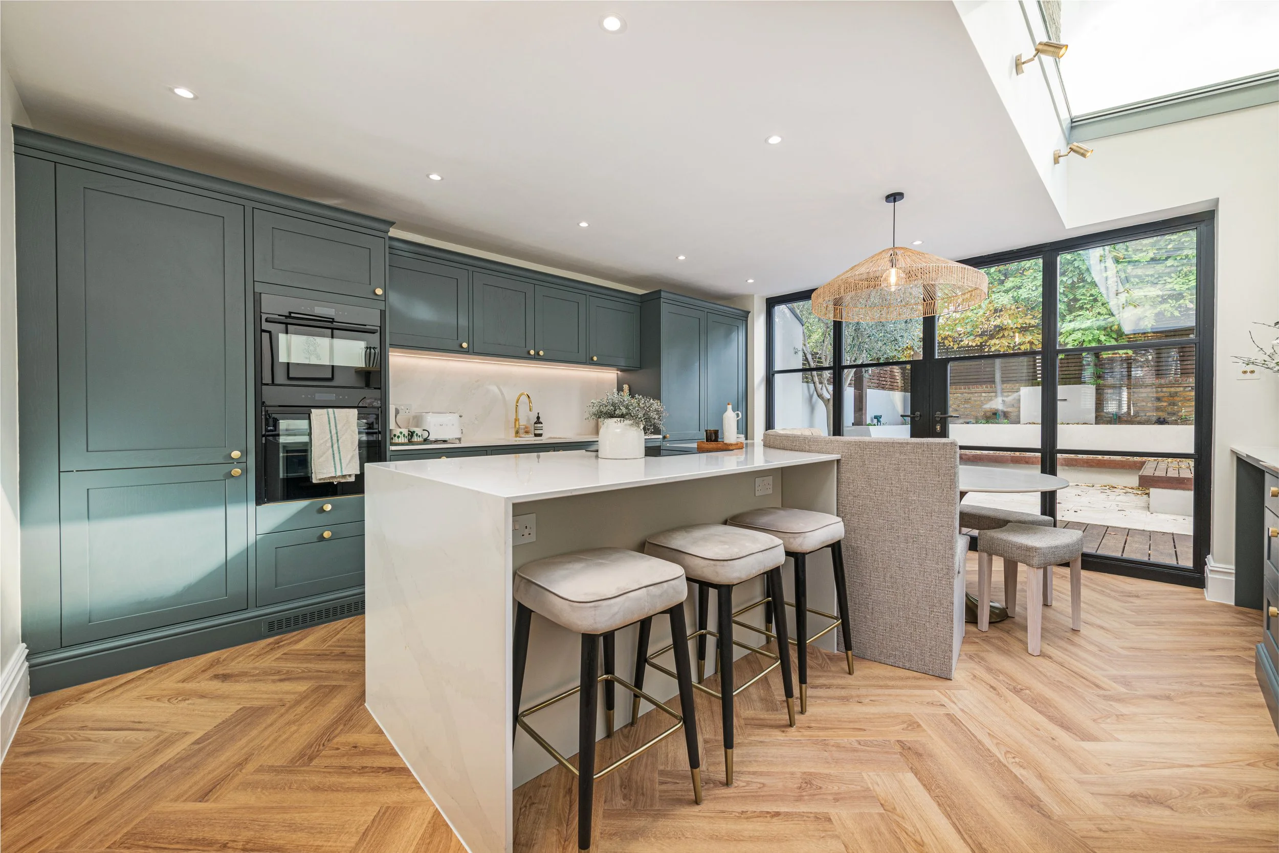 Modern open-concept kitchen with dark teal cabinets, white marble island, bar stools, a large window with a view of backyard, and hardwood flooring.