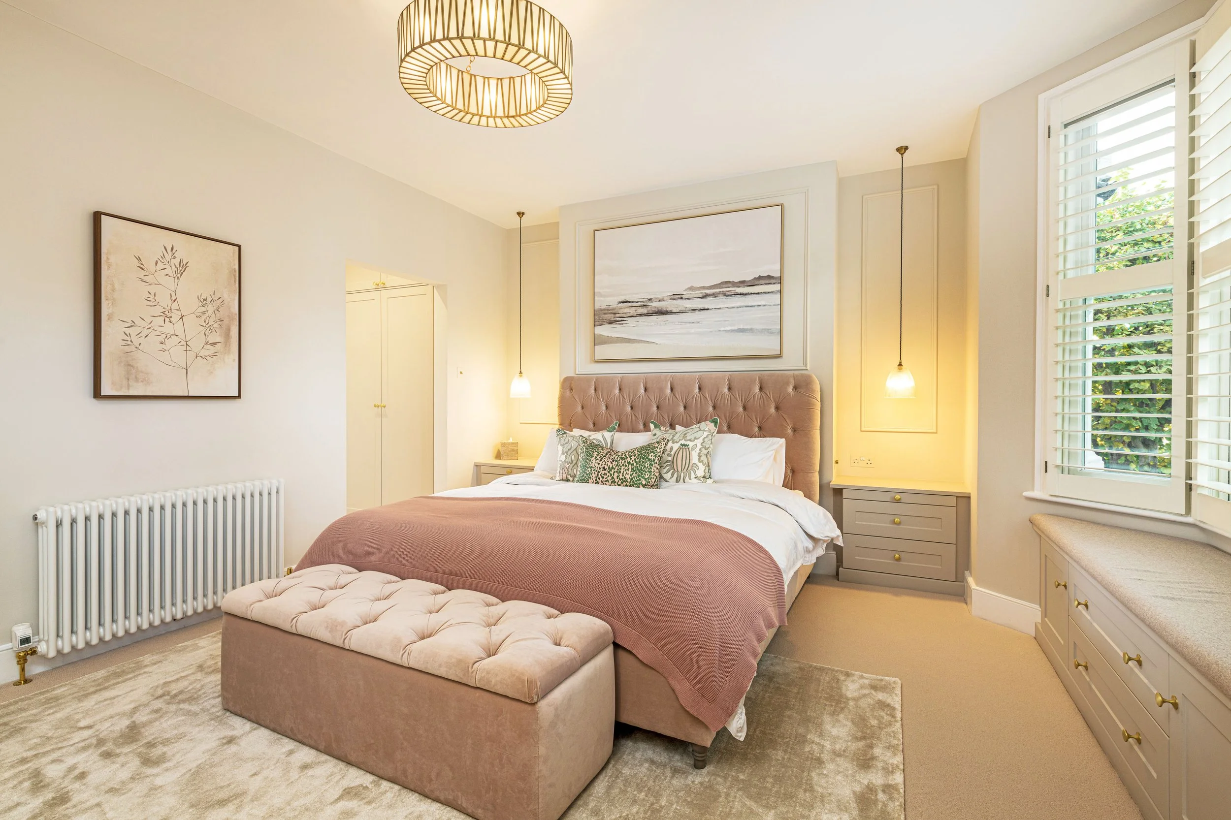 A cozy bedroom with a large bed featuring a pink headboard, patterned pillows, and a pink bedspread, flanked by two bedside tables with hanging pendant lights, large windows with white shutters, and artwork on the walls.