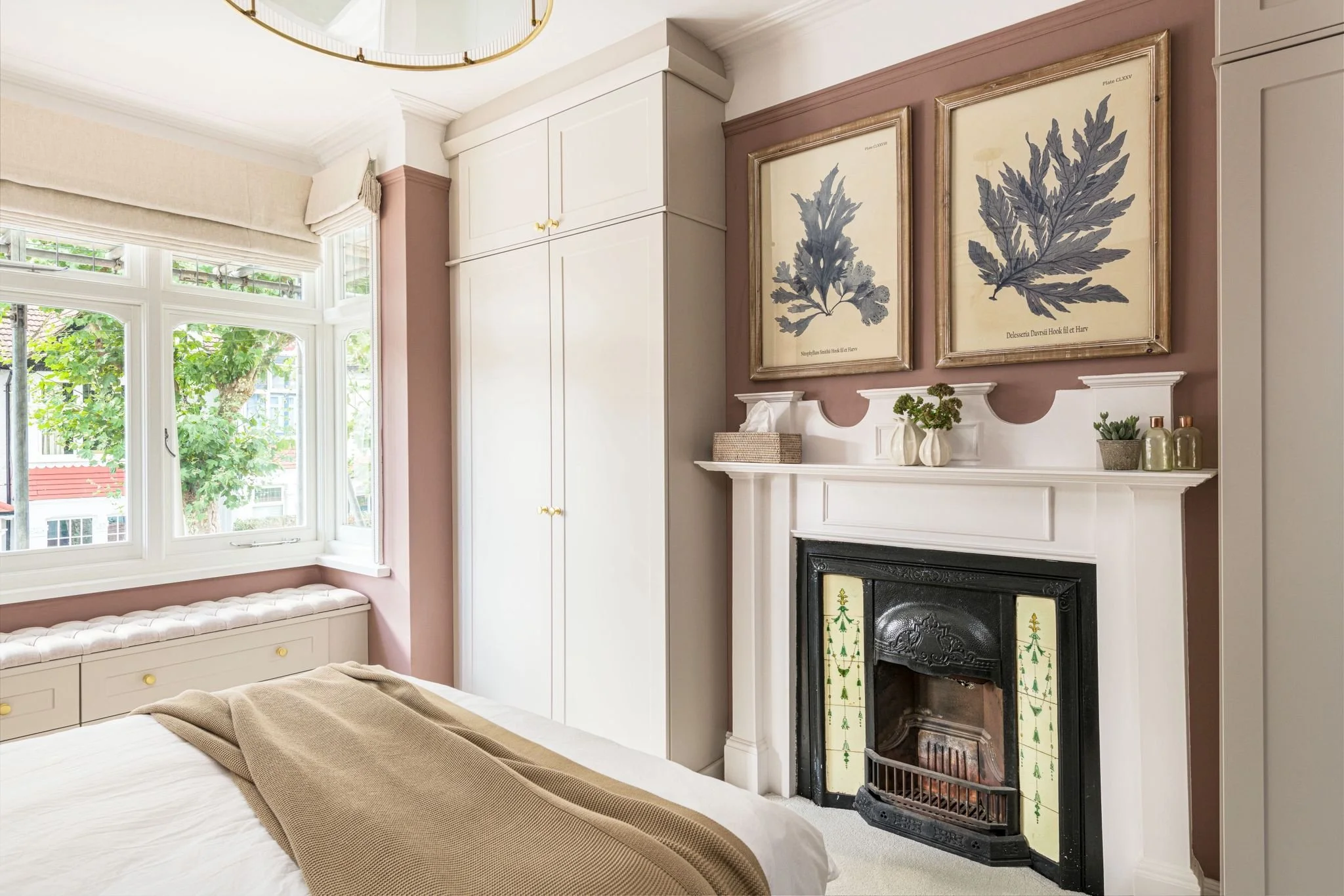 A cozy bedroom with a white fireplace mantel decorated with small vases and plants, a large window with white trim and beige curtains, and framed botanical prints on the pink-colored wall.
